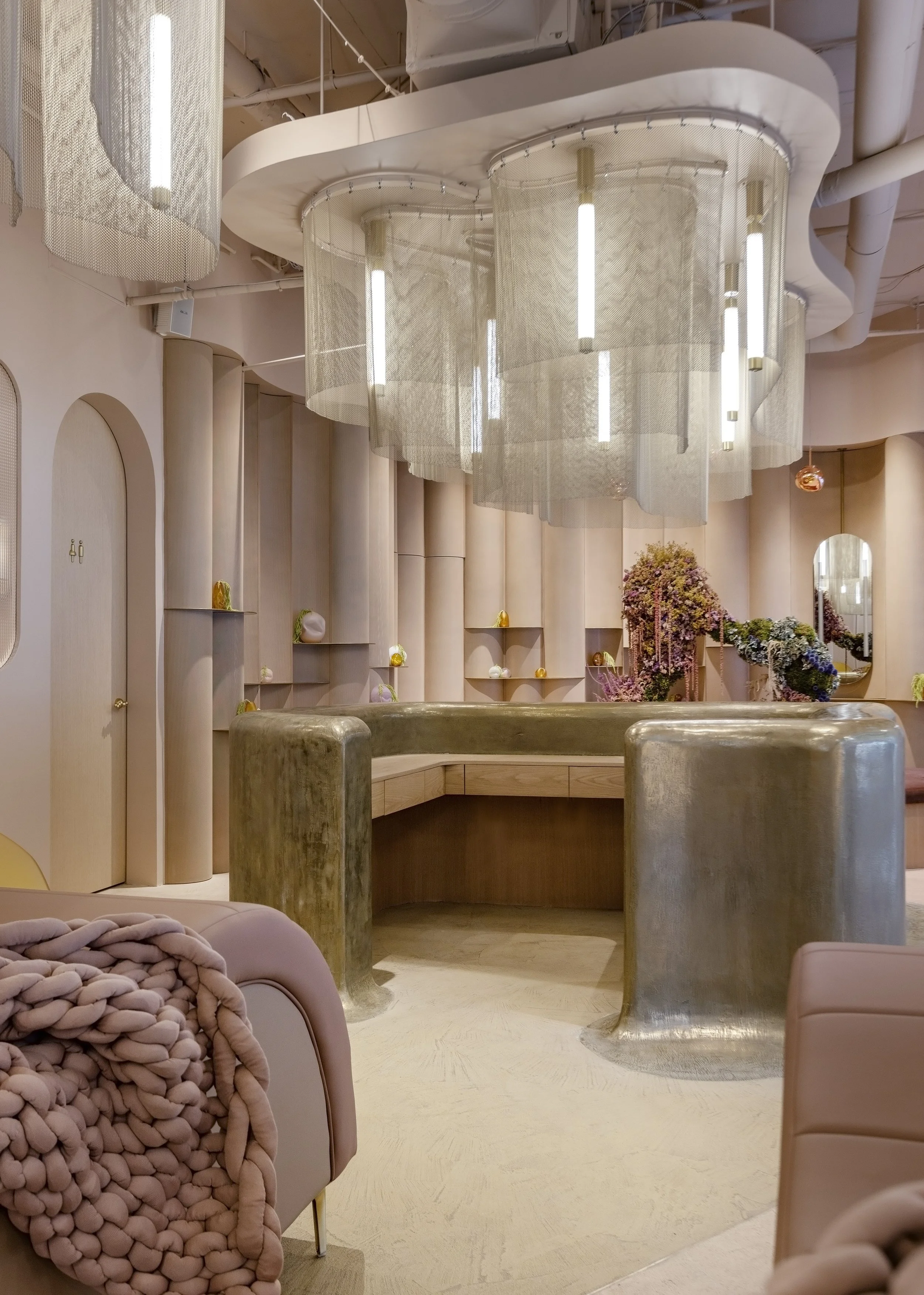Interior of a modern, stylish room with curved furniture, soft pastel colors, decorative vases, floral arrangements, and large hanging mesh panels with integrated lighting, creating an elegant and contemporary ambiance.