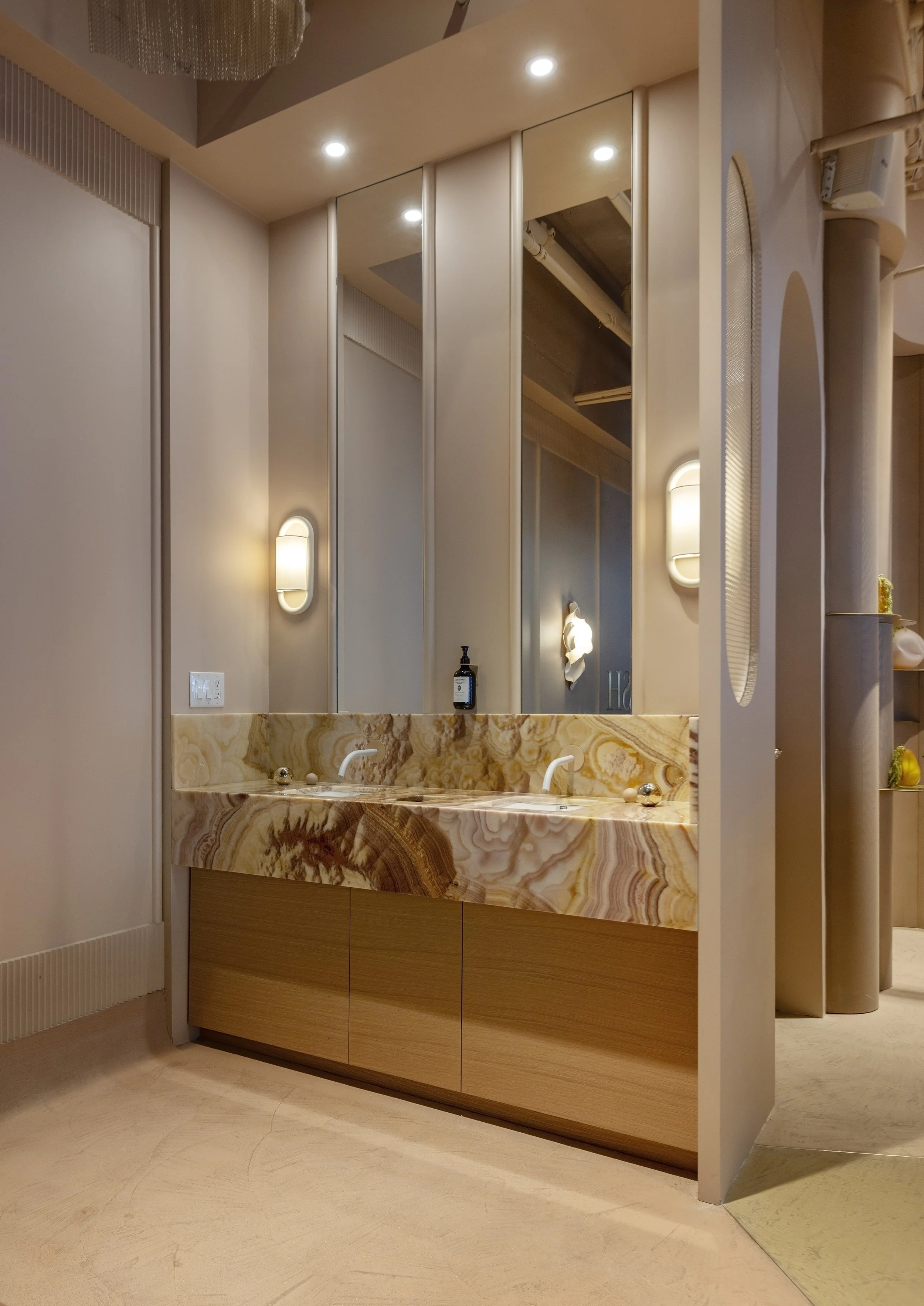 A modern bathroom vanity with a marble countertop and a built-in sink. There are three overhead mirrors, a soap dispenser, and two wall-mounted lights on either side of the mirrors. The vanity is made of wood with a curved front design.