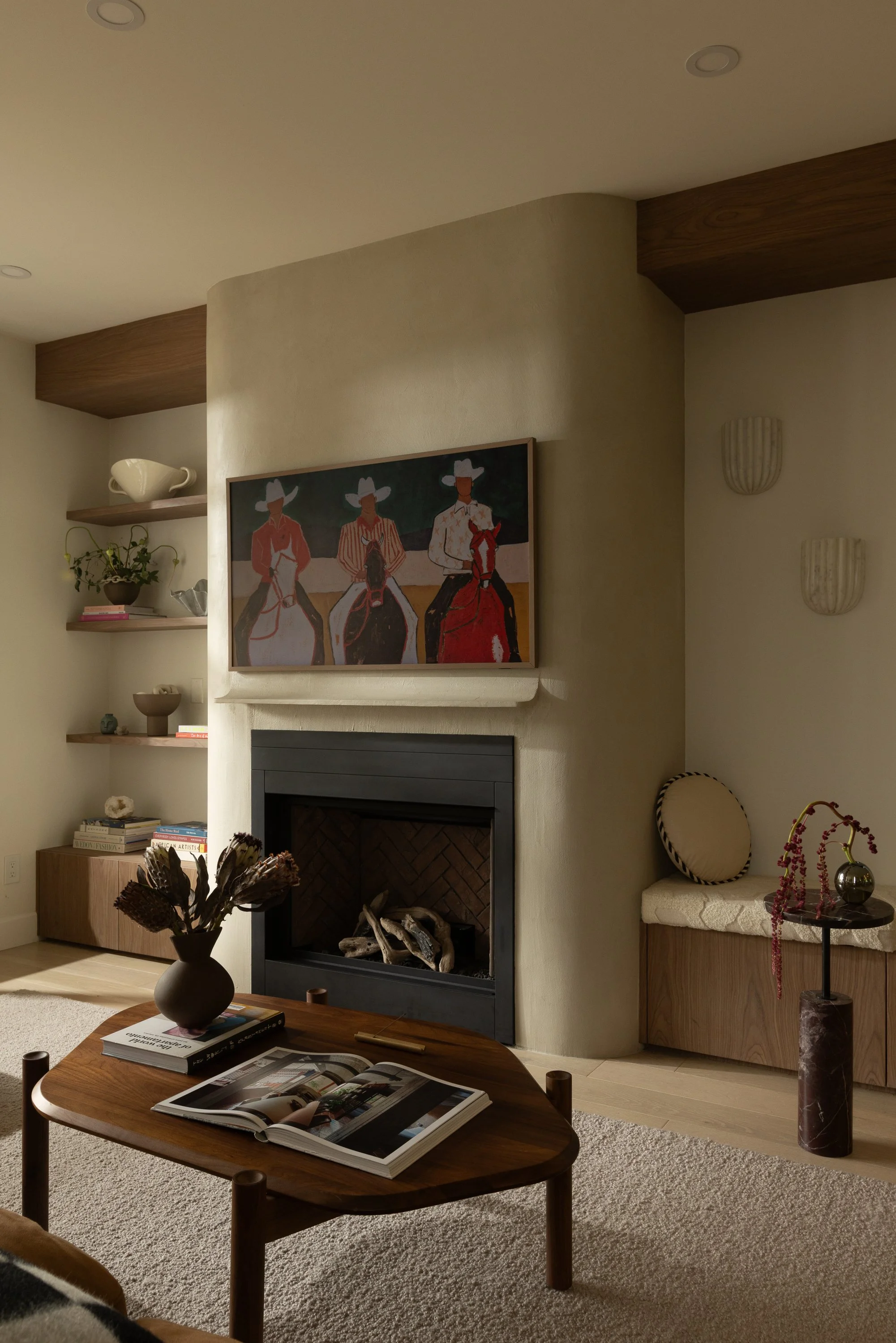 Living room with fireplace, framed artwork of three women in cowboy hats, decorative shelves, a vase with flowers on a wooden table, and a round upholstered chair next to a cabinet with a marble vase and pink beads.