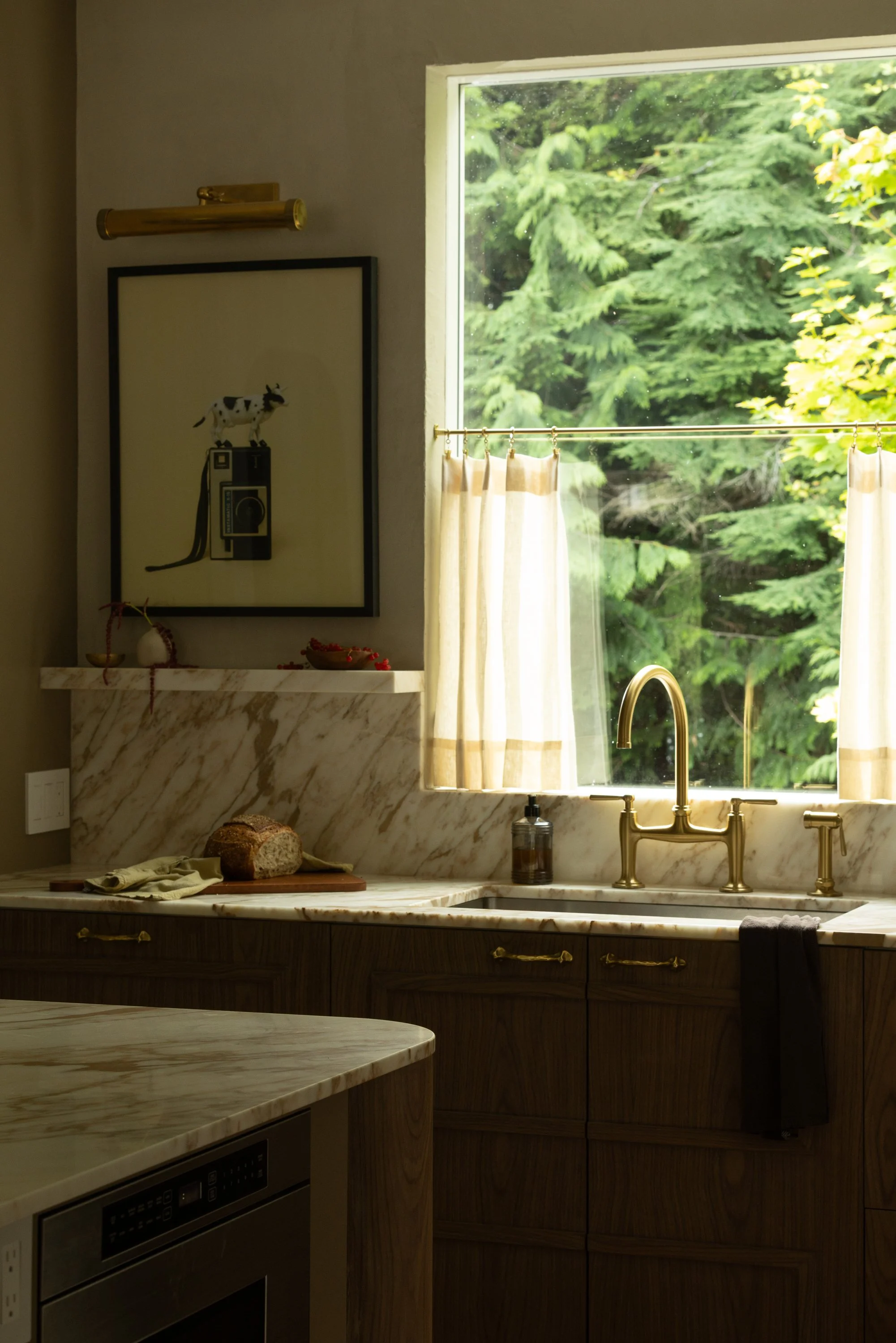 Kitchen scene with marble countertop, wooden cabinets, a gold faucet, a window with beige curtains showing green trees outside, a framed artwork of a cow on a camera, and a loaf of bread on a cutting board.