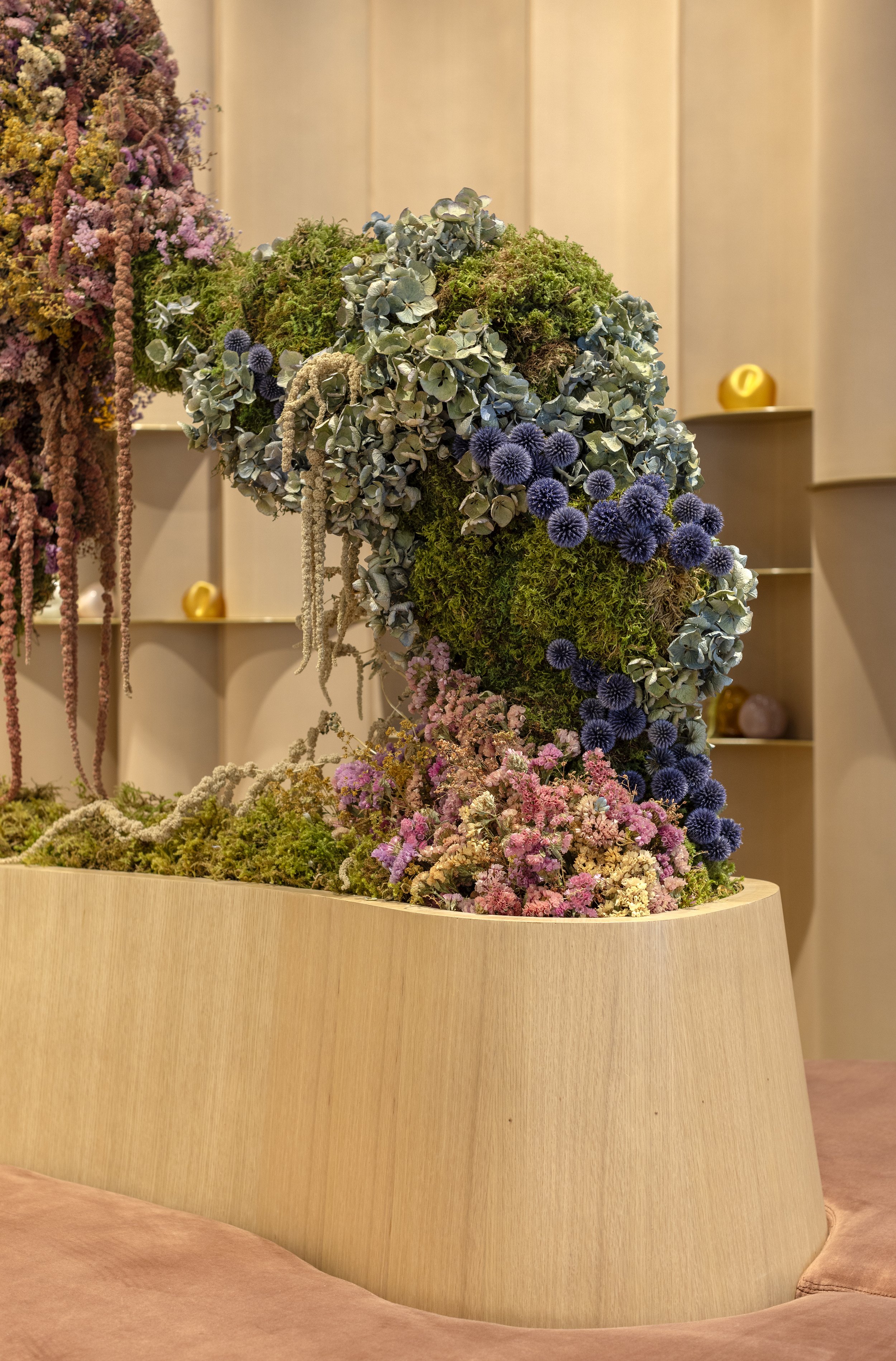 Decorative arrangement of various flowers and greenery on a wooden surface, with a background of soft beige paneling and small yellow decorative objects.