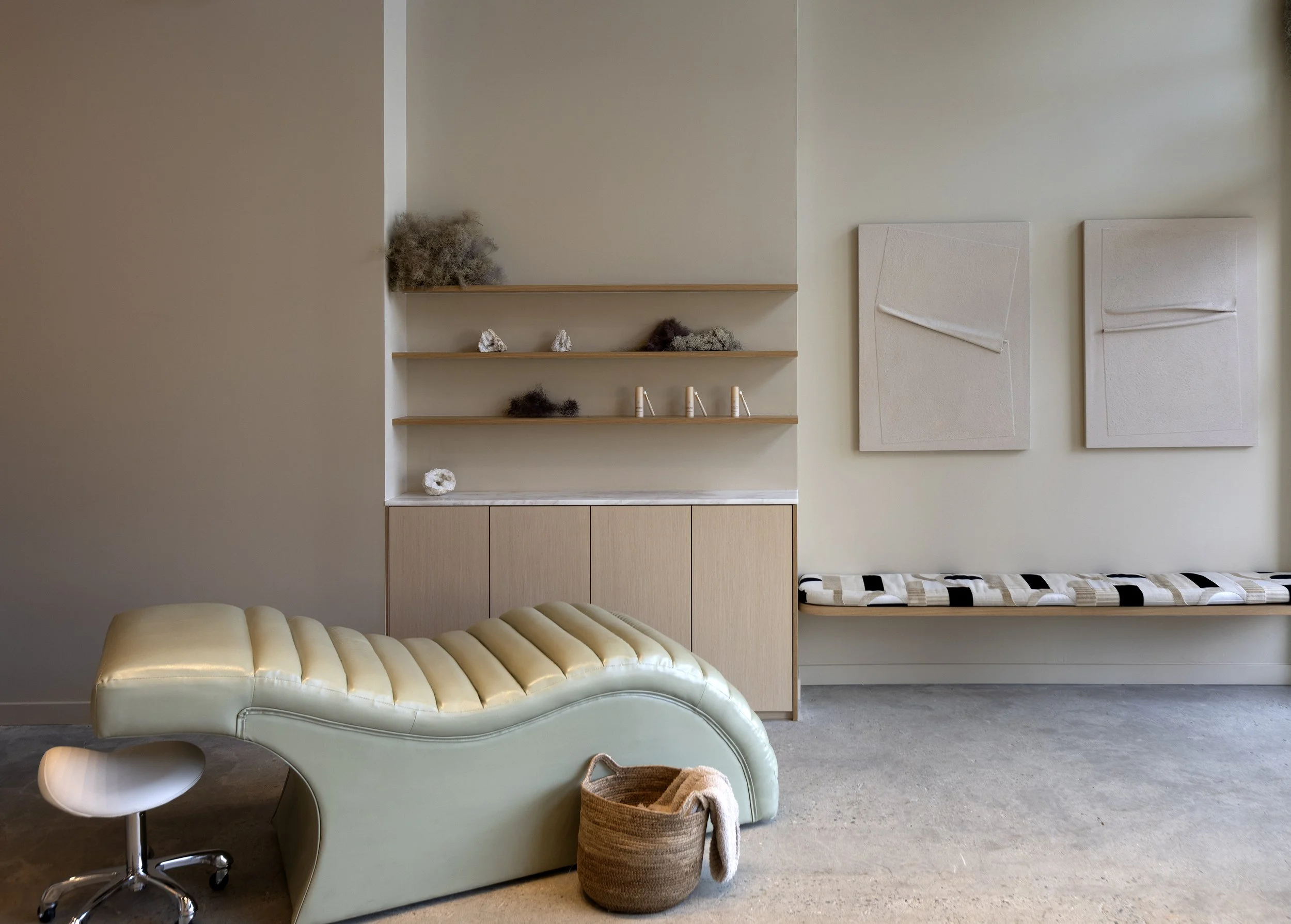 Interior of a room with beige walls, a light green curved sofa with tufted upholstery, a round glass table with a chrome stand, a woven basket with a towel inside, and wall-mounted shelves with decorative rocks and dried plants, along with abstract w