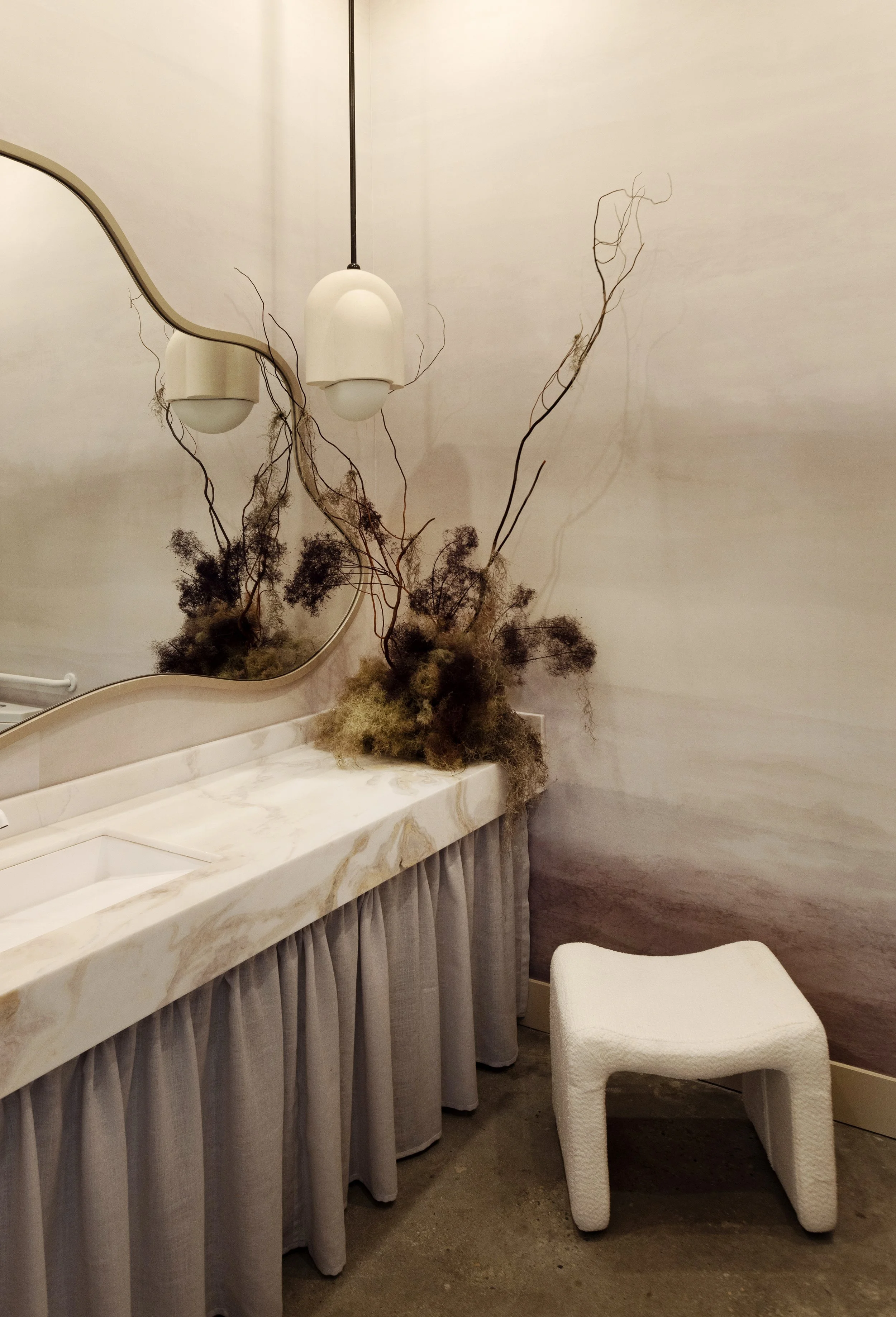 A bathroom or vanity area with a marble countertop, a mirror, a wall decoration resembling roots or branches, and a white upholstered chair.