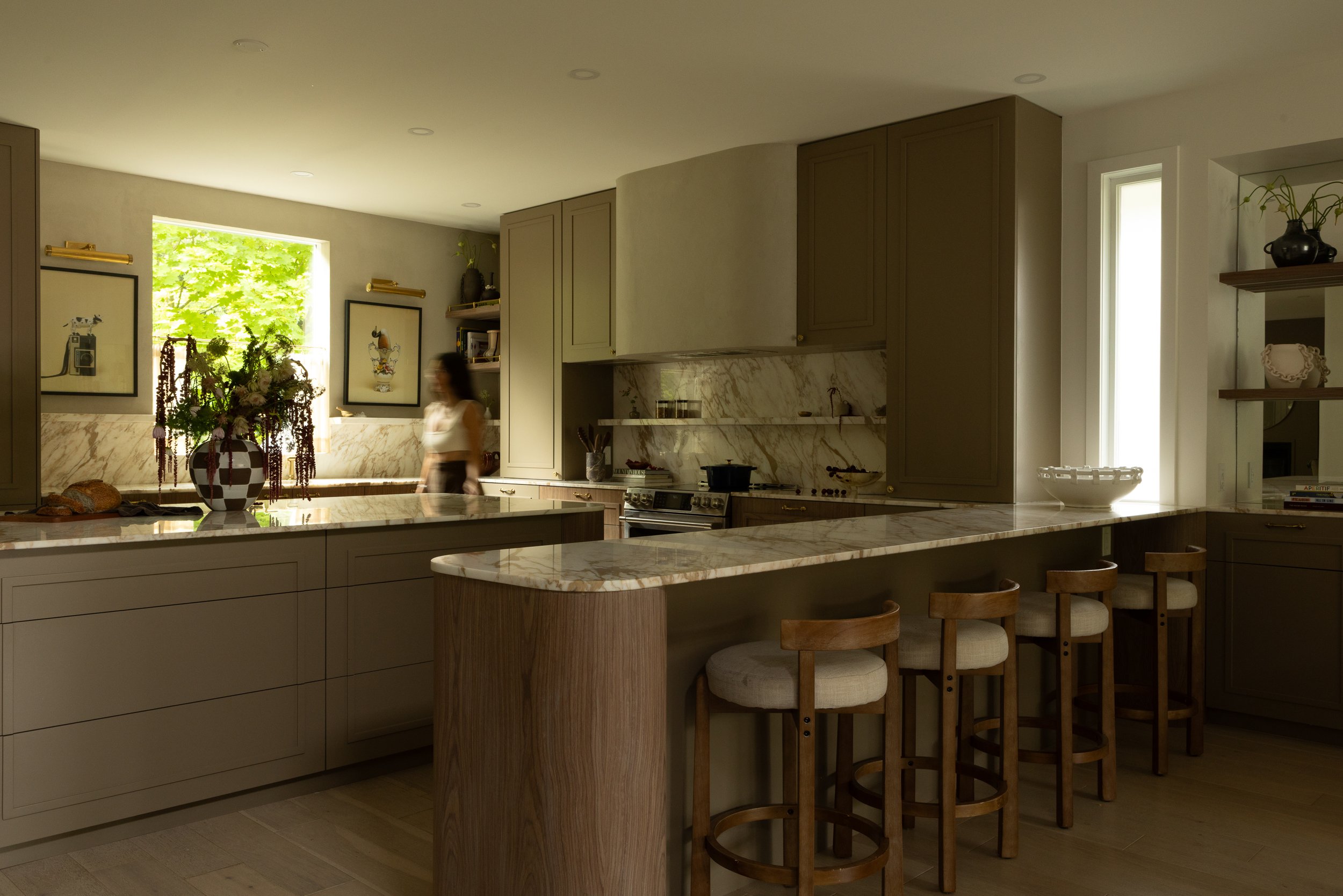 Modern kitchen with beige cabinets, marble countertops, and a breakfast bar with wooden stools. Sunlight coming through narrow windows. Decor includes framed art, a vase with greenery, and kitchen utensils.