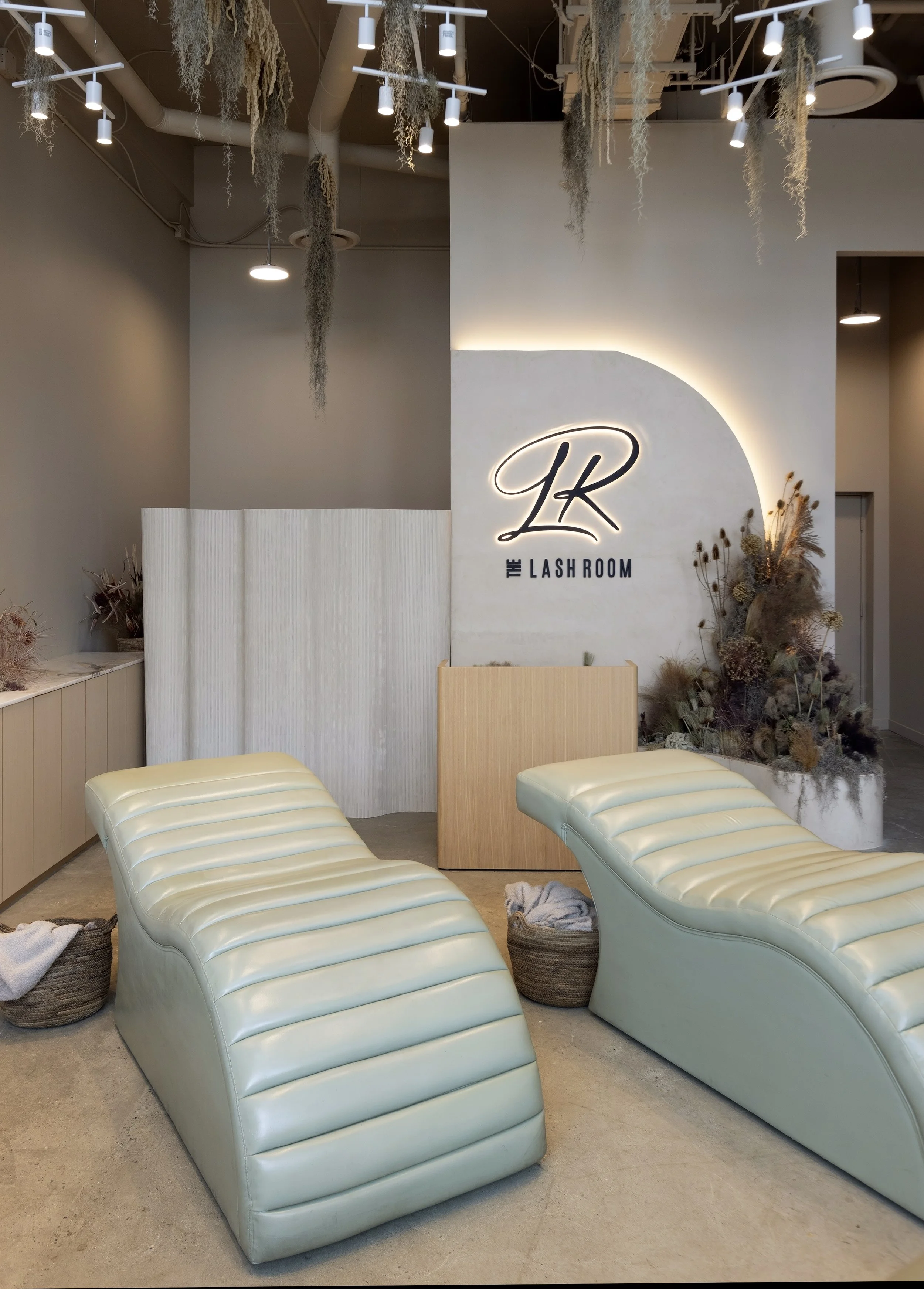 Interior of a modern lash salon with two cream-colored lounge chairs, basket storage, and a decorative wall with a logo reading 'The Lash Room' surrounded by dried flowers and plants.