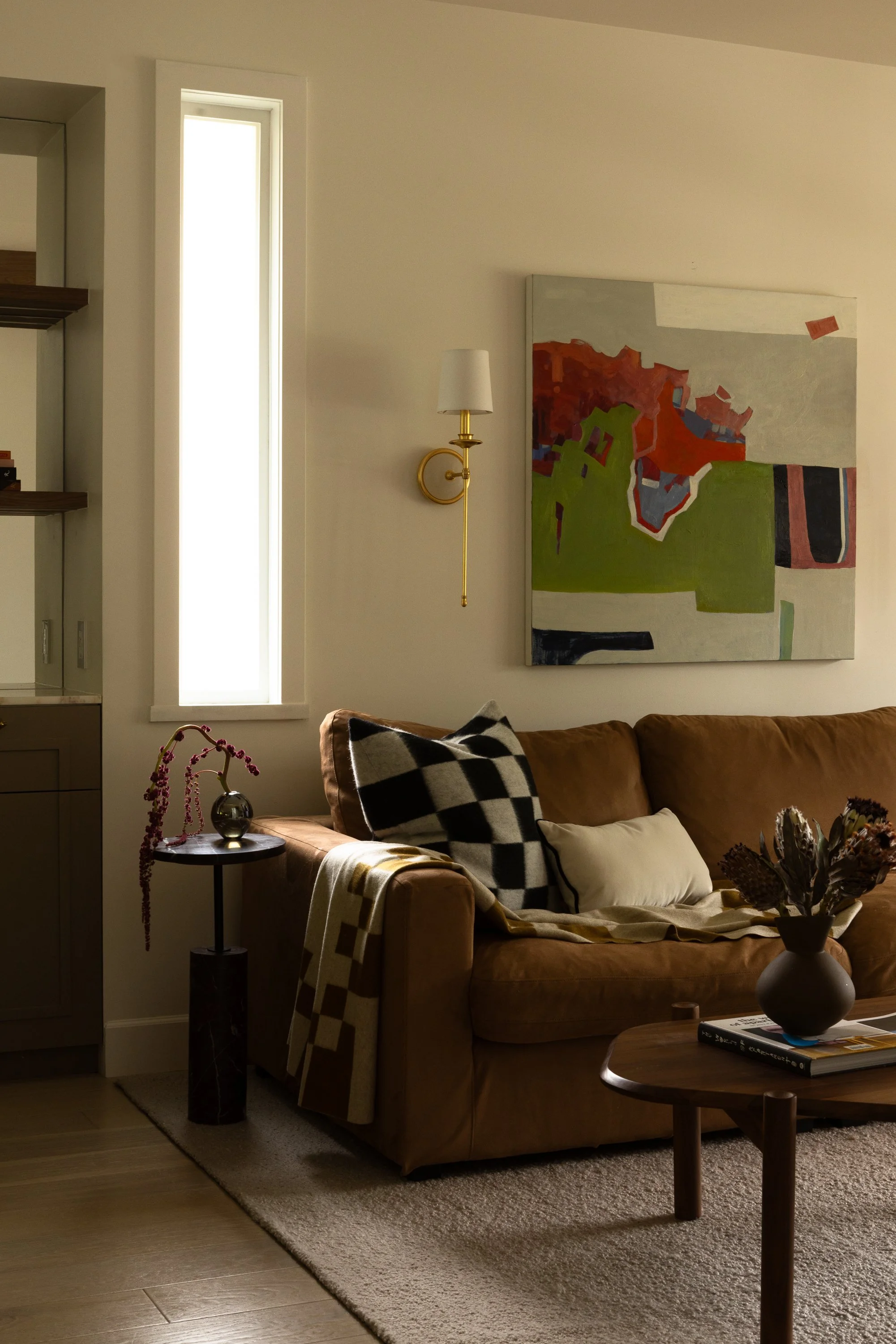 A cozy living room with a tan sofa adorned with black and white checkered and white pillows. A small round side table with a vase of pink decorative branches and a throw blanket. An abstract painting in red, green, and dark tones hangs on the wall. A wall-mounted light fixture is above the sofa, and a tall narrow window is next to it. The room features a beige carpet and wooden flooring.