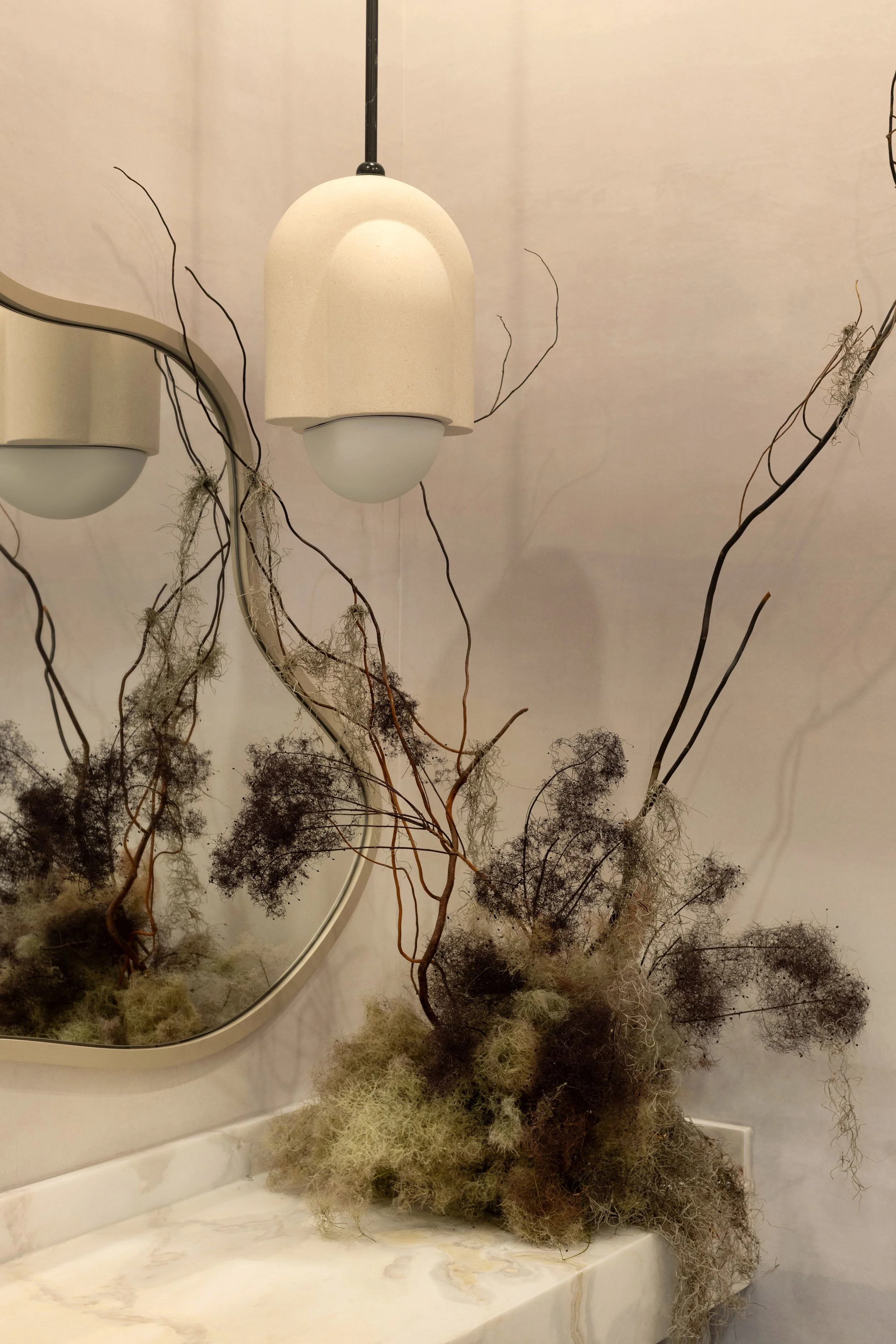 A modern hanging light fixture with a beige shade and white glass bottom, reflected in an oval mirror, surrounded by dried, wispy branches and moss on a marble countertop.