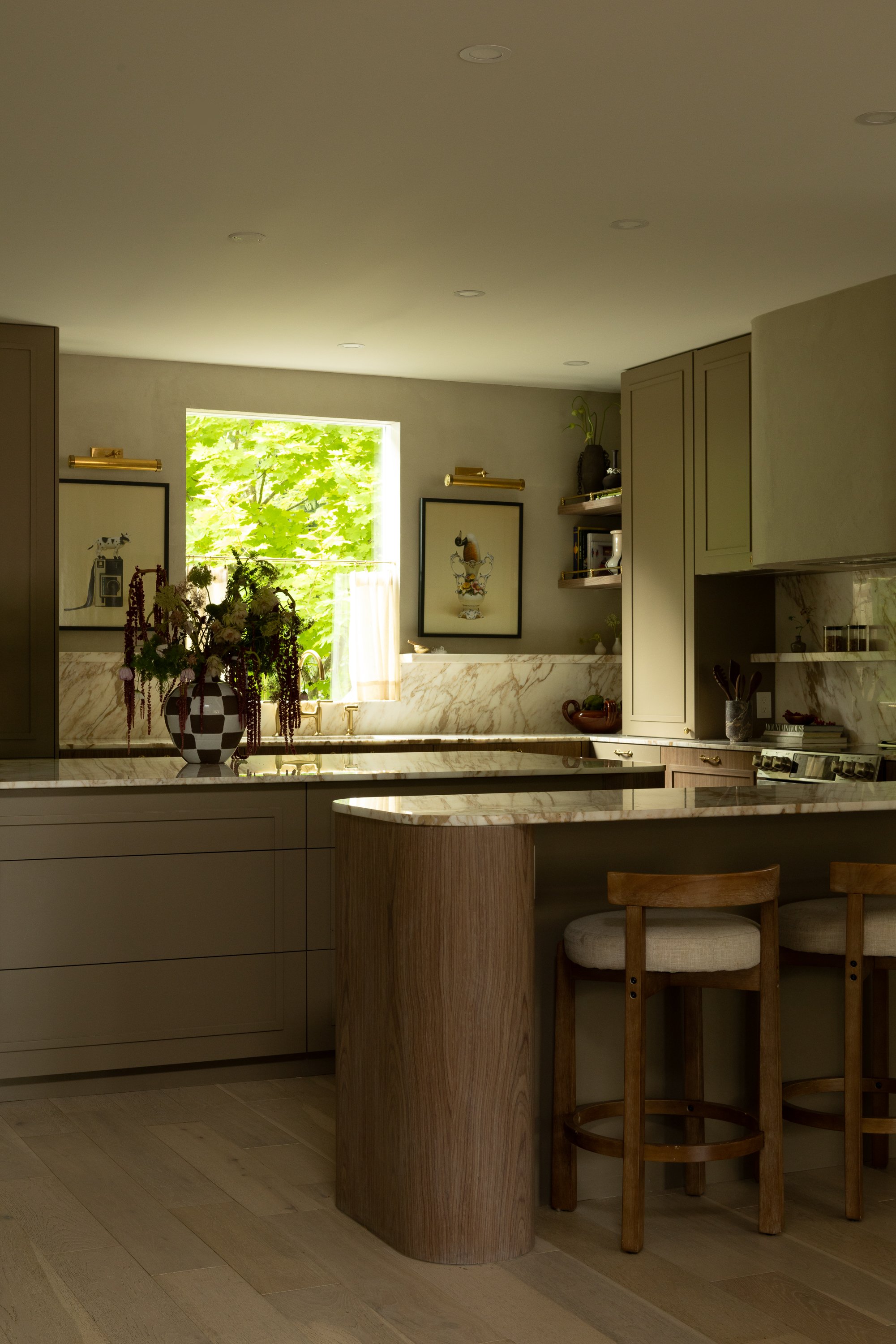 A modern kitchen with a marble countertop, a vase with flowers, framed artwork on the wall, beige cabinets, and a window with green trees outside. Two wooden chairs are at the kitchen island.