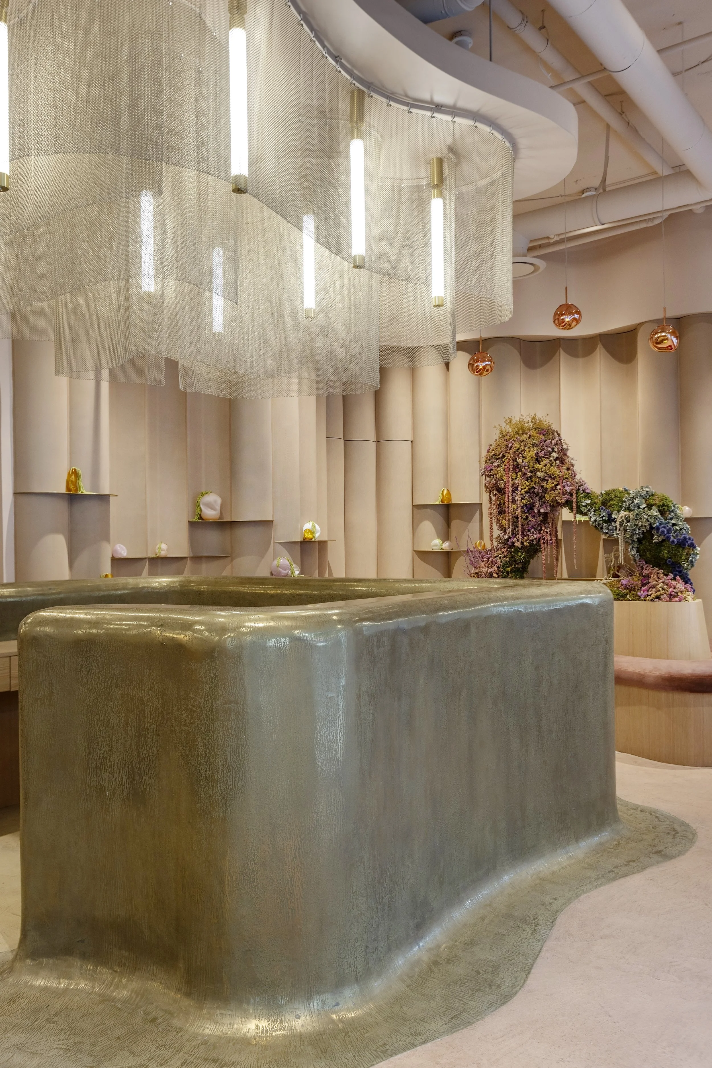 A modern interior space with a metallic curved reception desk in the foreground, decorative objects on shelves along the wall, and a floral arrangement with pink and purple flowers. The ceiling has exposed white pipes and hanging orange spherical light fixtures, with a large light-colored fabric fixture hanging from the ceiling.