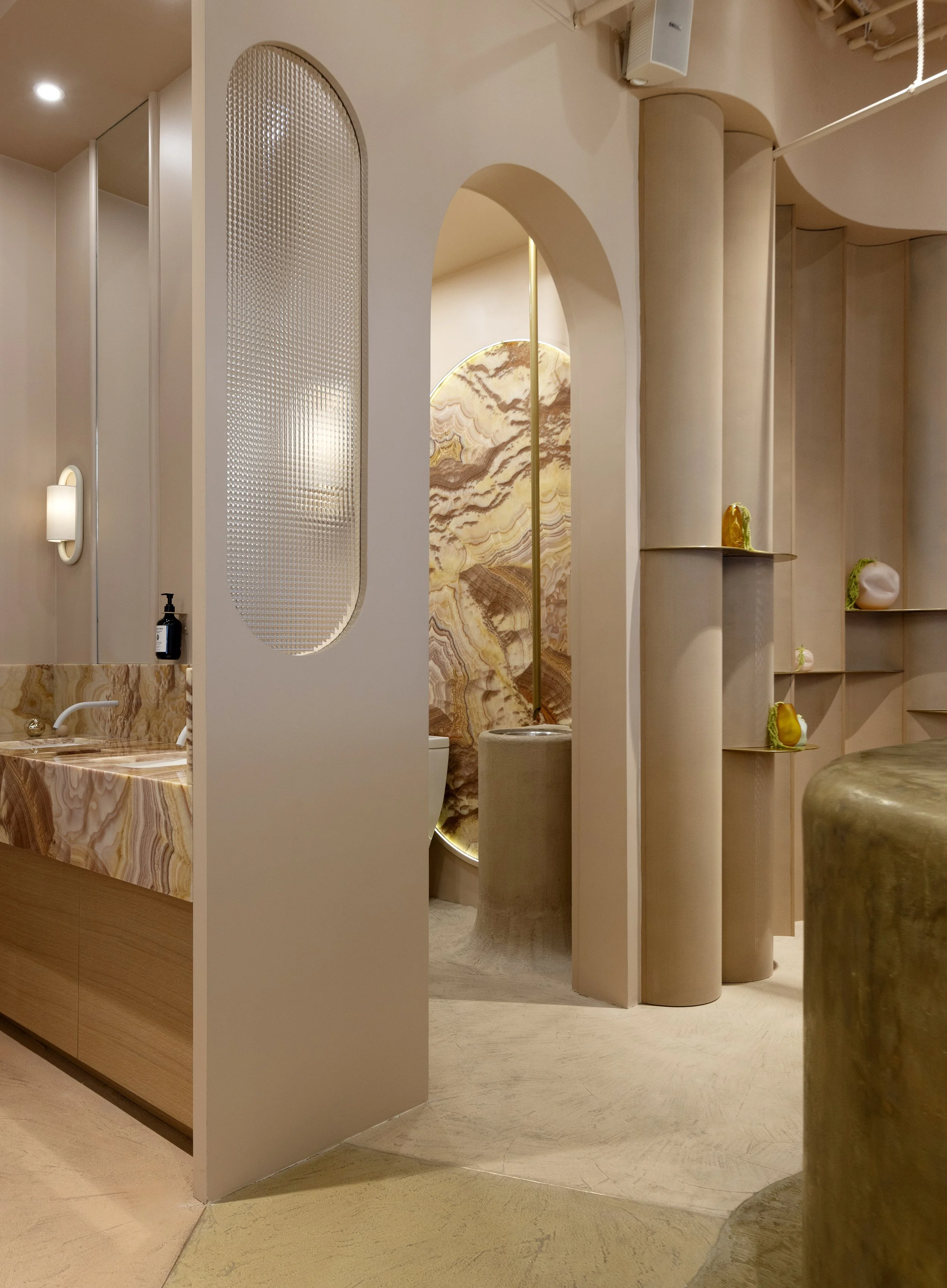 A modern interior space with curved architectural elements, soft lighting, and decorative objects on shelves. There is a partition with textured glass, a marble-patterned wall, and a mix of warm neutral tones.