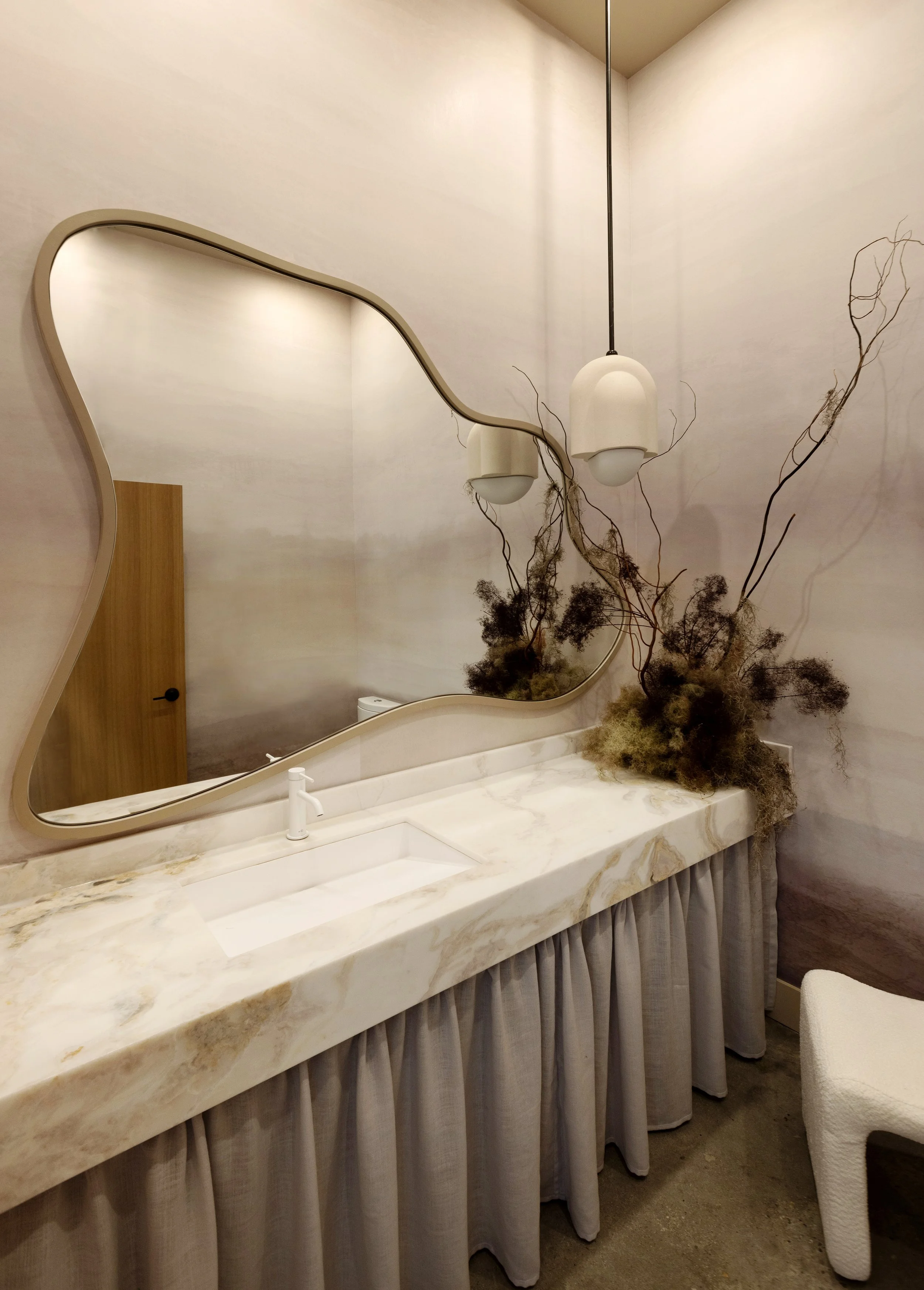 Elegant bathroom with a marble countertop, an oval mirror with a beige frame, hanging pendant lights, and dried floral arrangement near the sink.