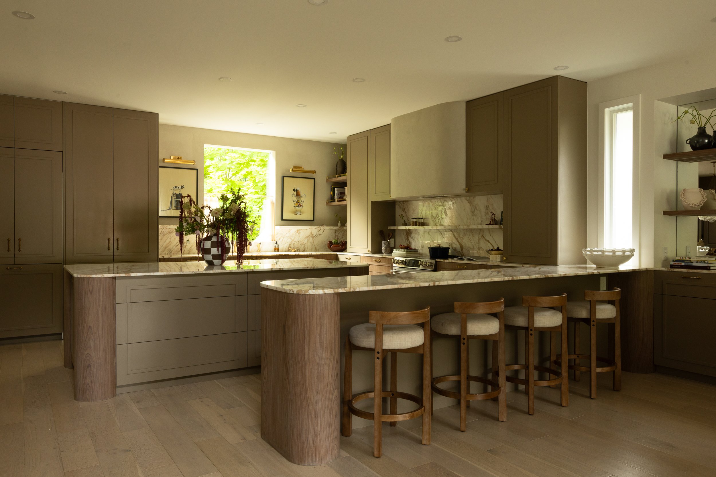 A modern kitchen with beige cabinets, a marble countertop, and a kitchen island with four wooden bar stools. Two framed pictures hang on the wall by a window with green trees outside, and decorative items are placed on open shelves and countertops.