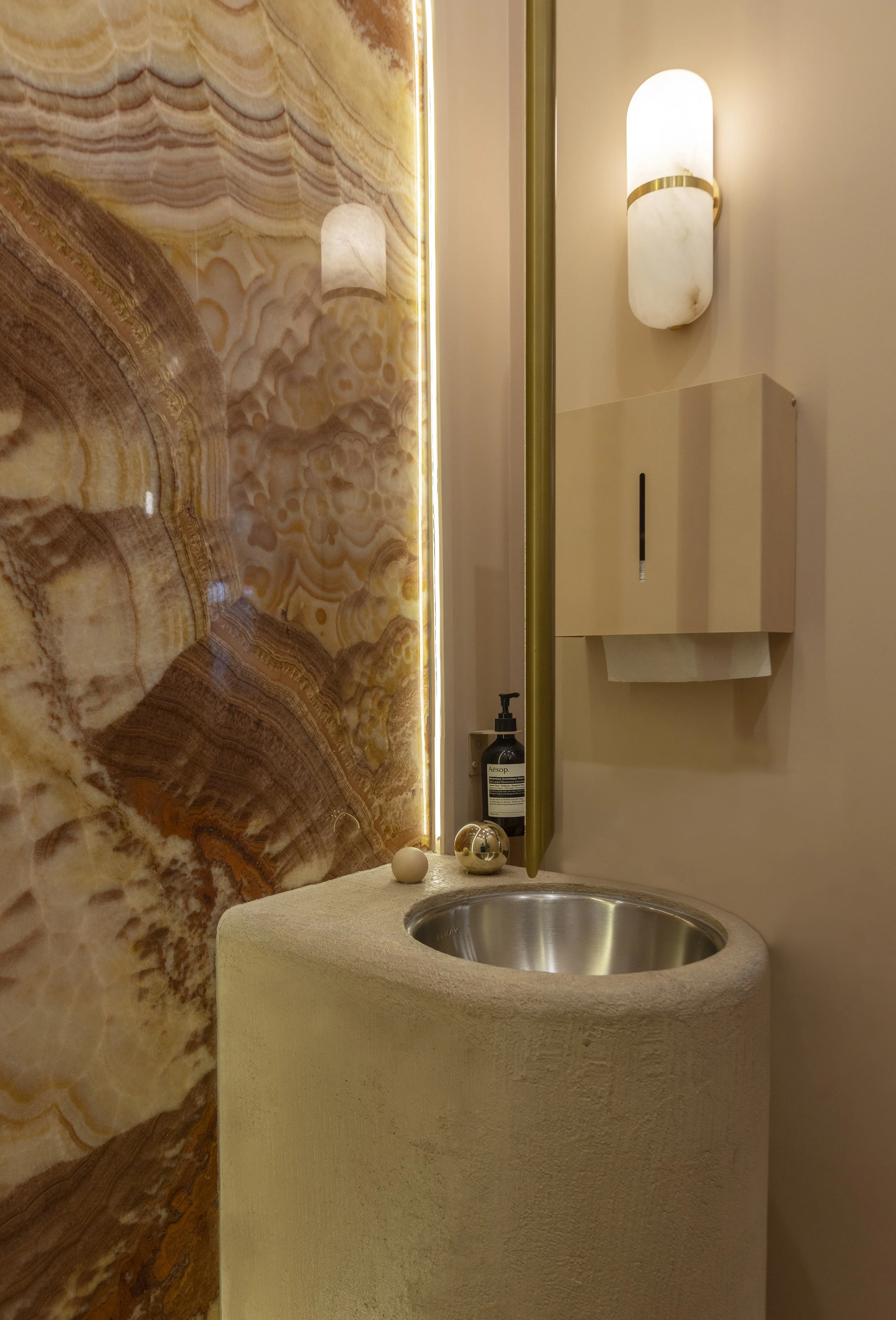A bathroom sink with a textured beige countertop, a round stainless steel basin, decorative objects including a spherical sculpture, a small ball, a soap dispenser, a wall-mounted light fixture, a backlit onyx or marble wall, and a beige toilet paper holder.