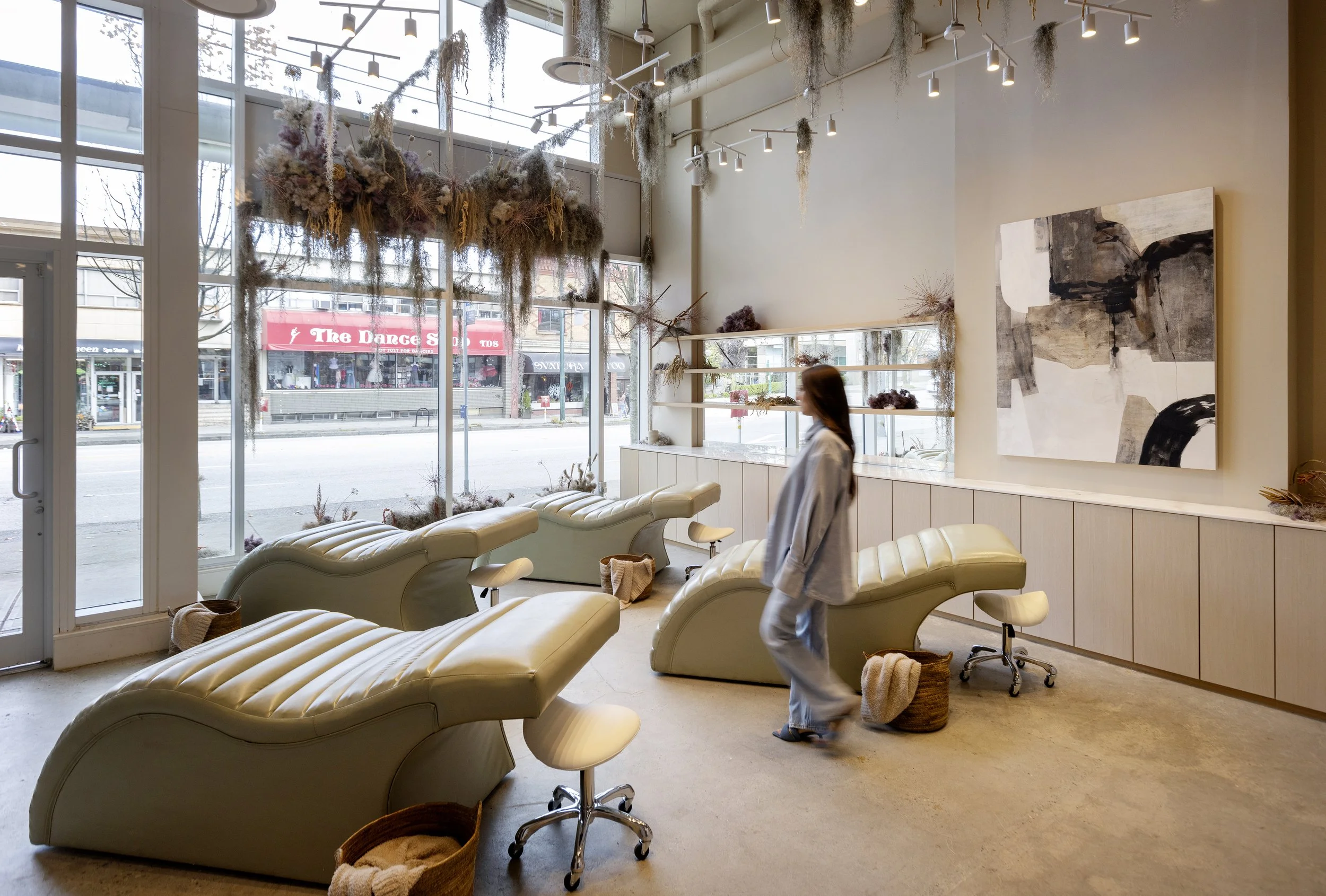 The photo shows the inside of a modern salon or waiting area with cream-colored leather chairs, a large window, wall art, and decorative dried plants hanging from the ceiling.