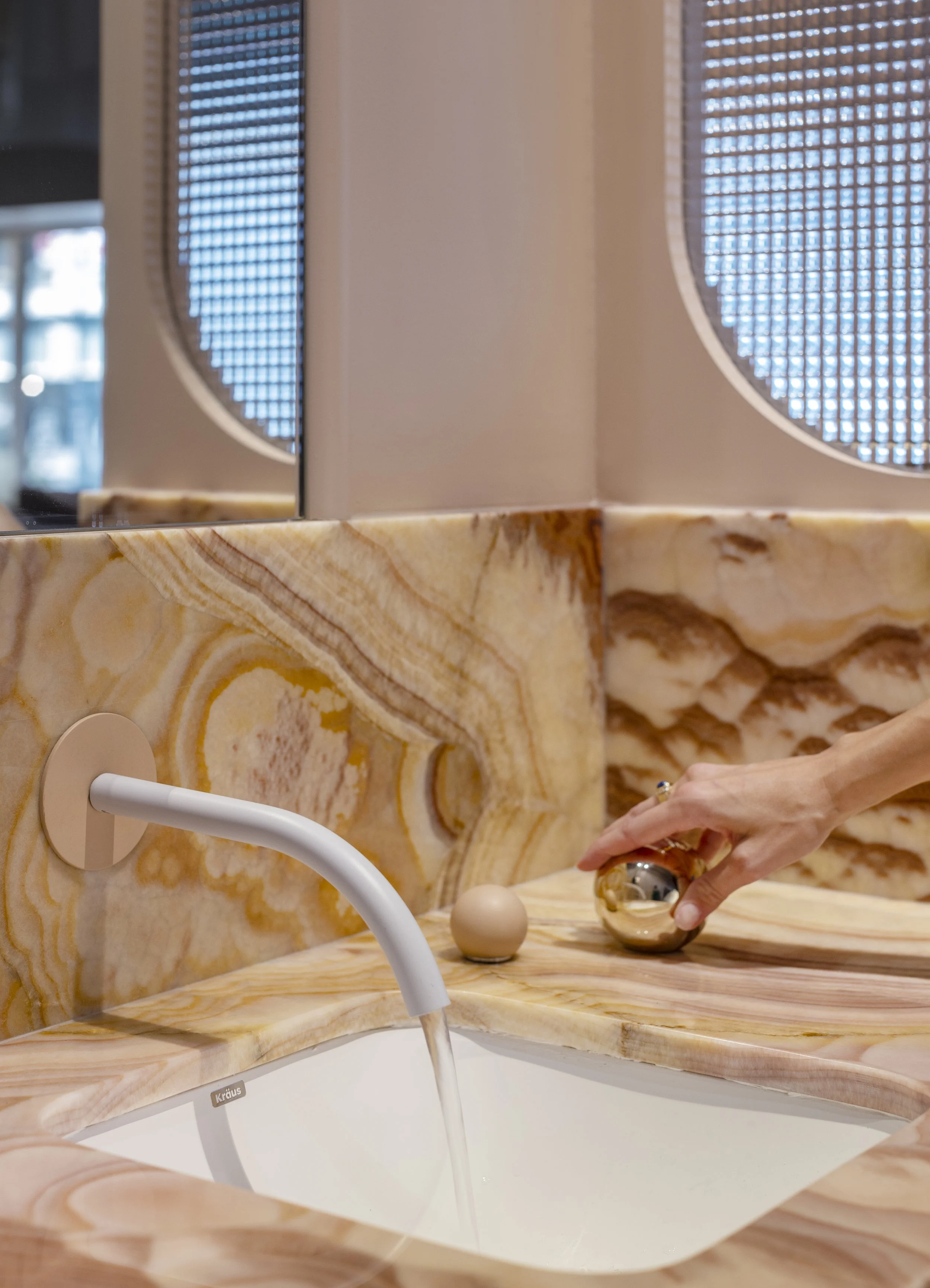 Close-up of a person's hand turning a shiny silver faucet handle above a white sink, with a beige and brown veined marble countertop. In the background, there is a wall with a large round decorative mirror and a window with a grid pattern.