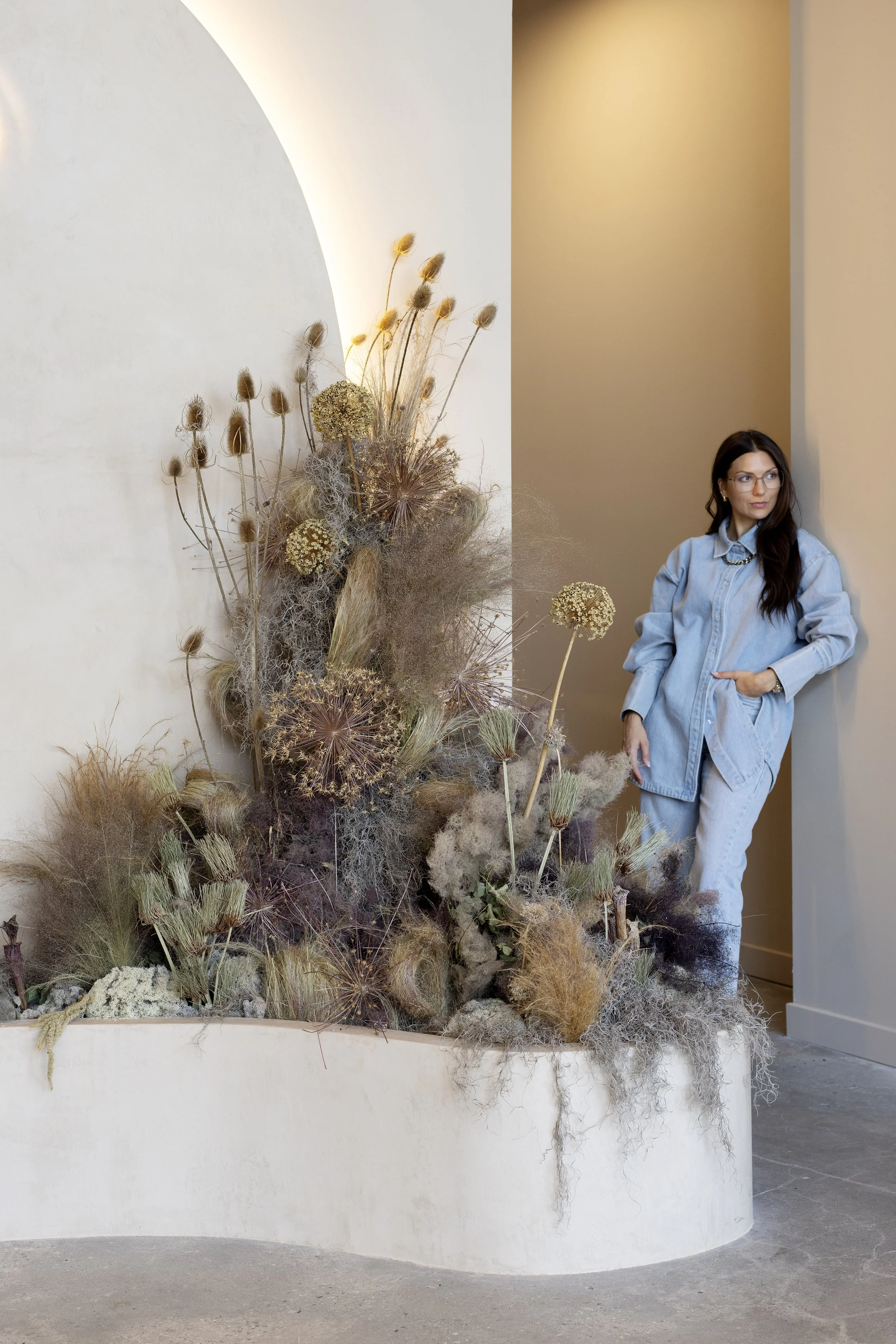 A woman with long dark hair, wearing glasses and a light blue oversized shirt and matching pants, standing beside an art installation of dried plants and flowers against a plain wall.