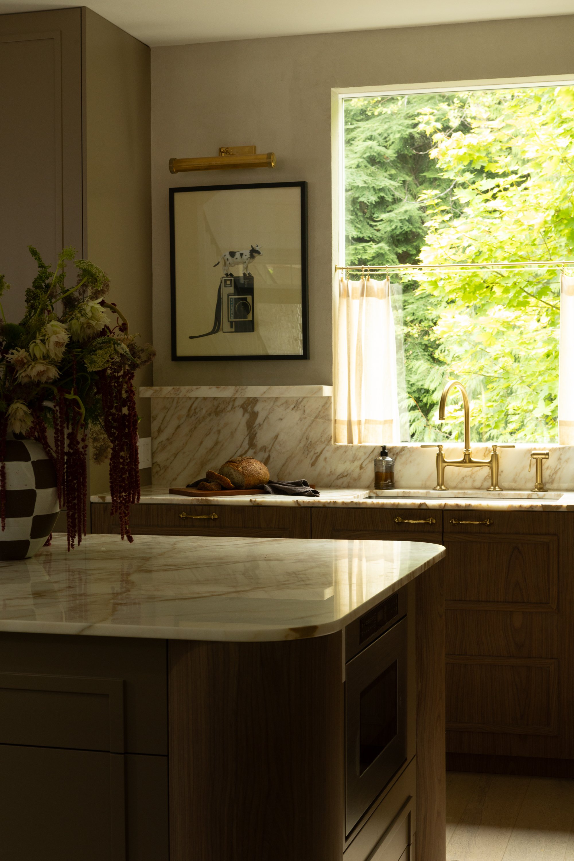 A kitchen with a marble countertop, wooden cabinets, a window showing green trees outside, a golden faucet, a floral arrangement, and a picture of a cow on the wall.