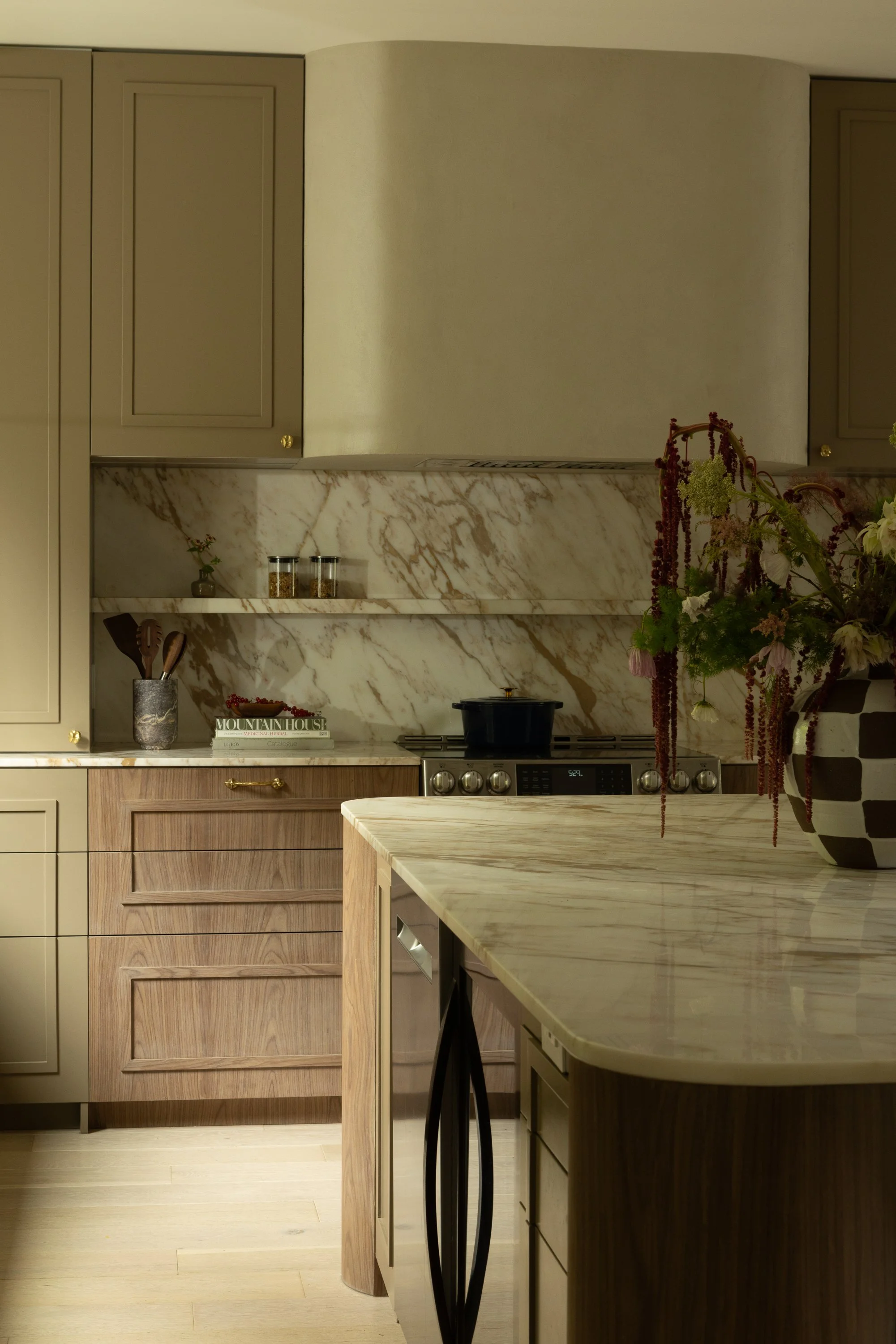 A modern kitchen with beige and wooden cabinetry, a marble backsplash, a marble countertop, and a bouquet of flowers in a black vase on the counter.