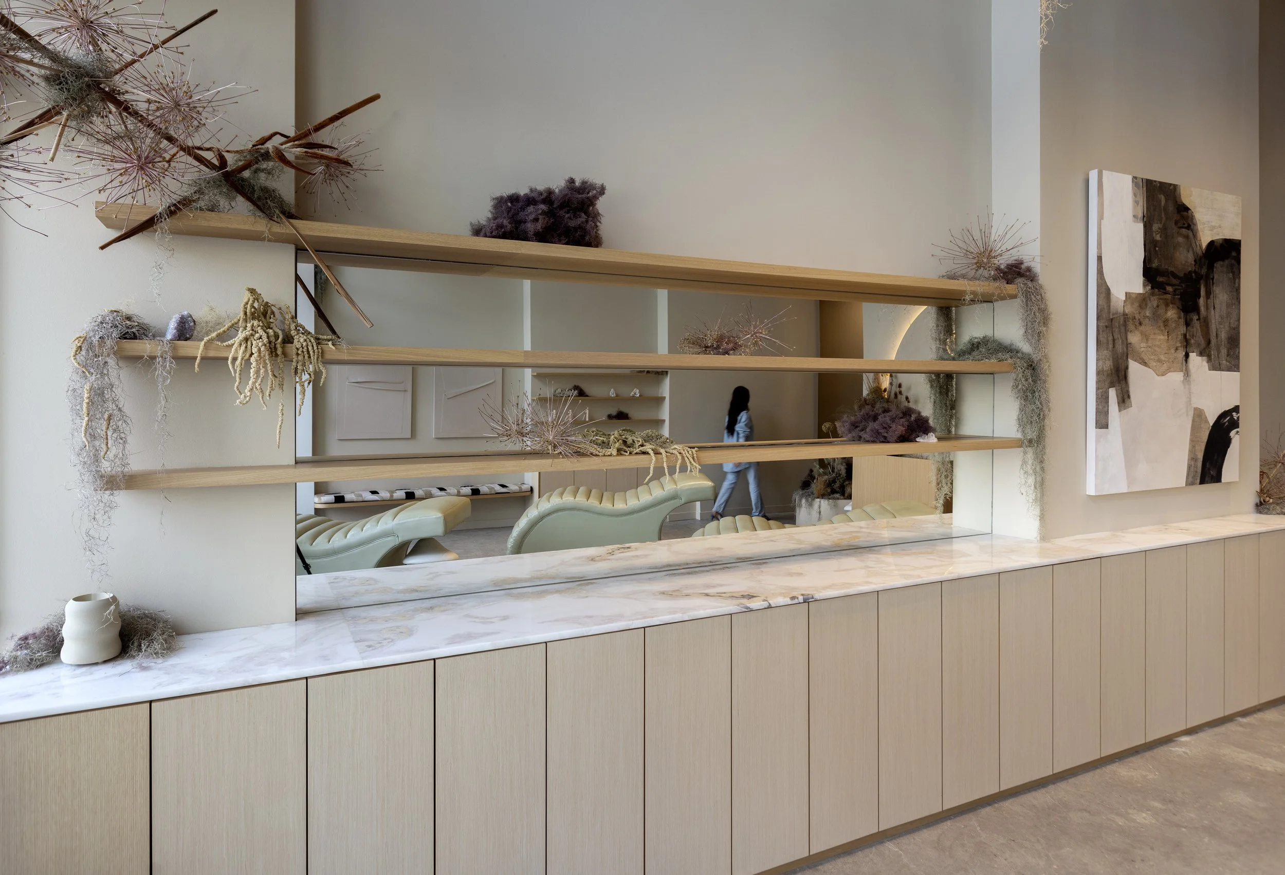 A modern interior space with wooden shelves decorated with dried plants and accessories, a marble counter, a cream-colored chair, and abstract artwork on the wall.