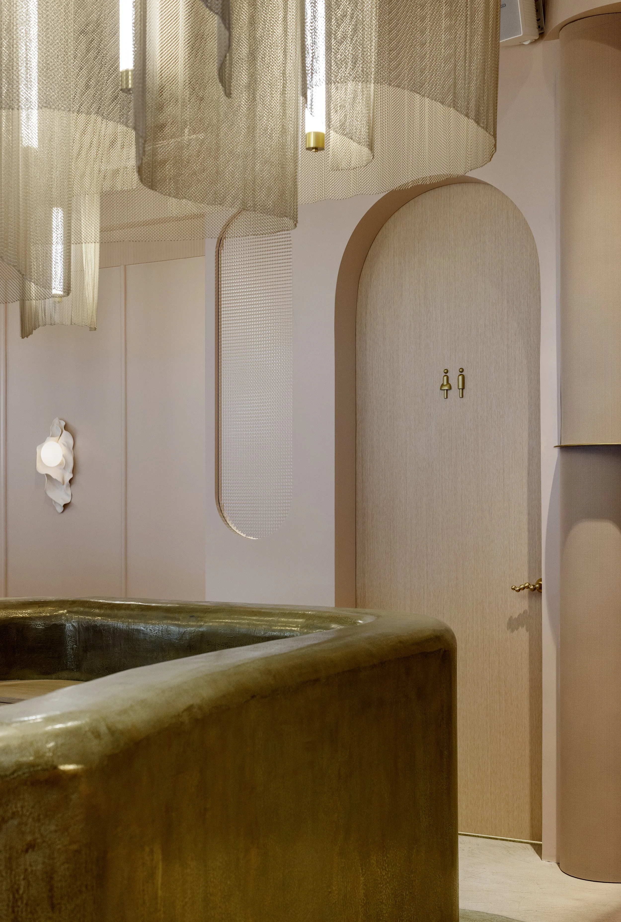 A beige wall with a decorative archway featuring small gold figure door handles, beige textured curtains hanging from a rod, and part of a stone bathtub in the bottom foreground.