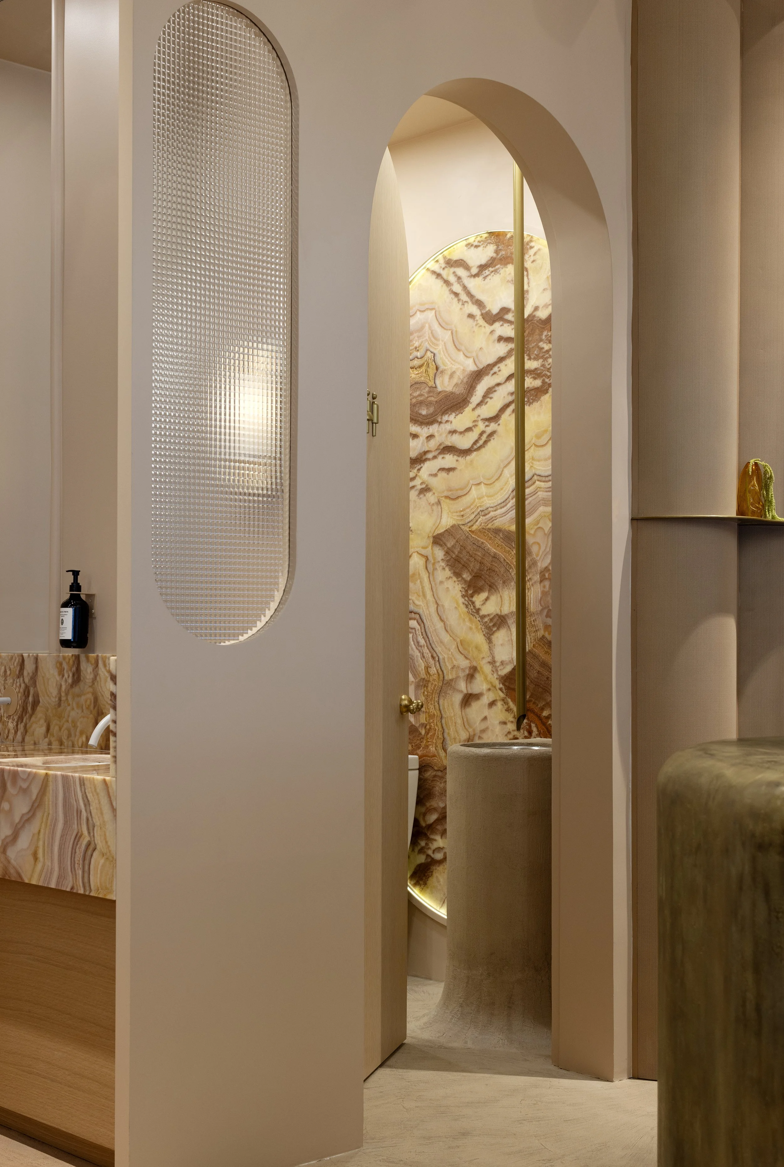Interior view of a bathroom with a marble onyx wall, a door with a curved top, and a stool. A curtain is partially visible on the right side, and a small shelf with a decorative item is on the wall.