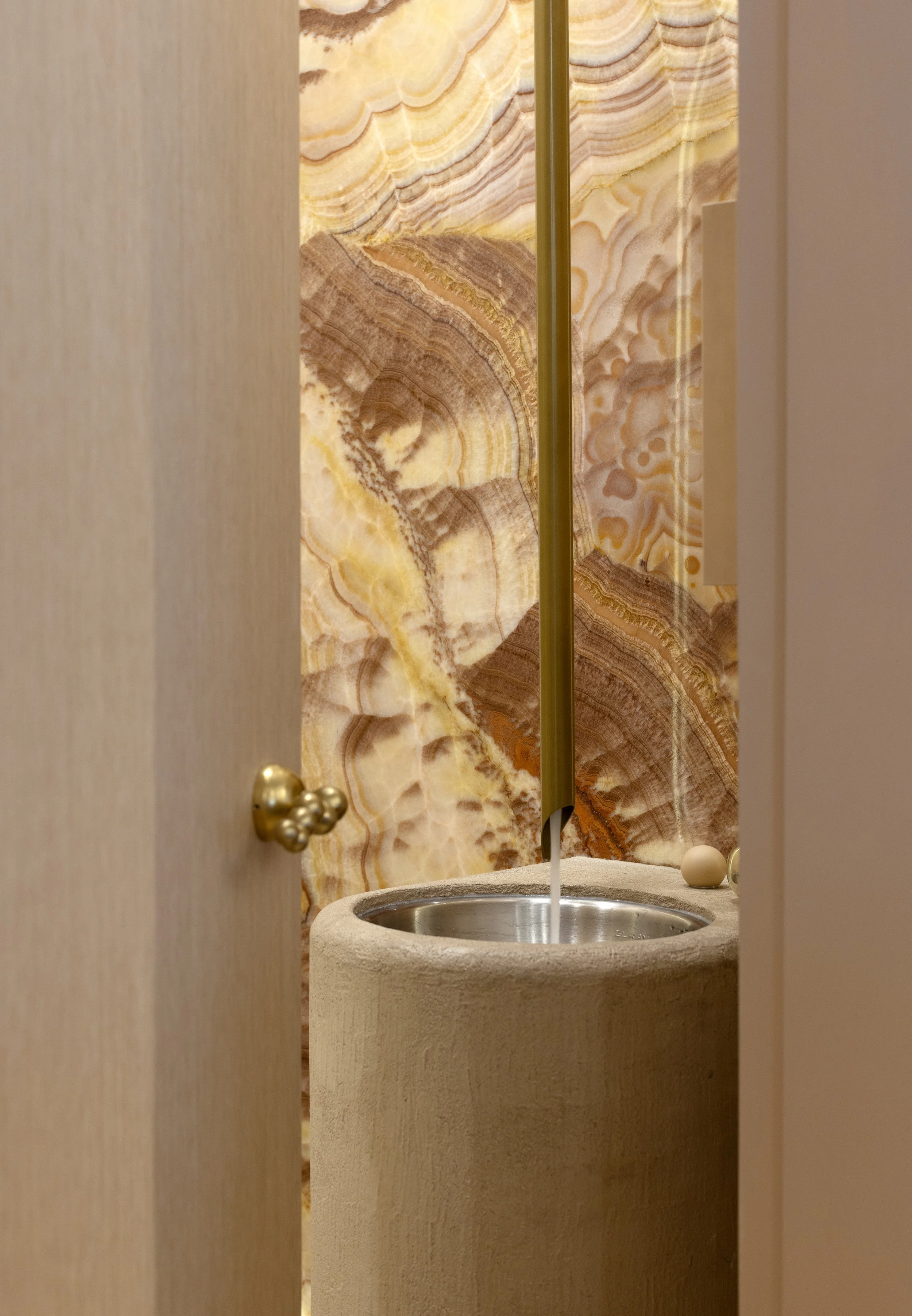 A bathroom view through a partially open door showing a beige stone sink with water running into it, a gold-colored faucet, and a marble-patterned wall with warm colors in the background.