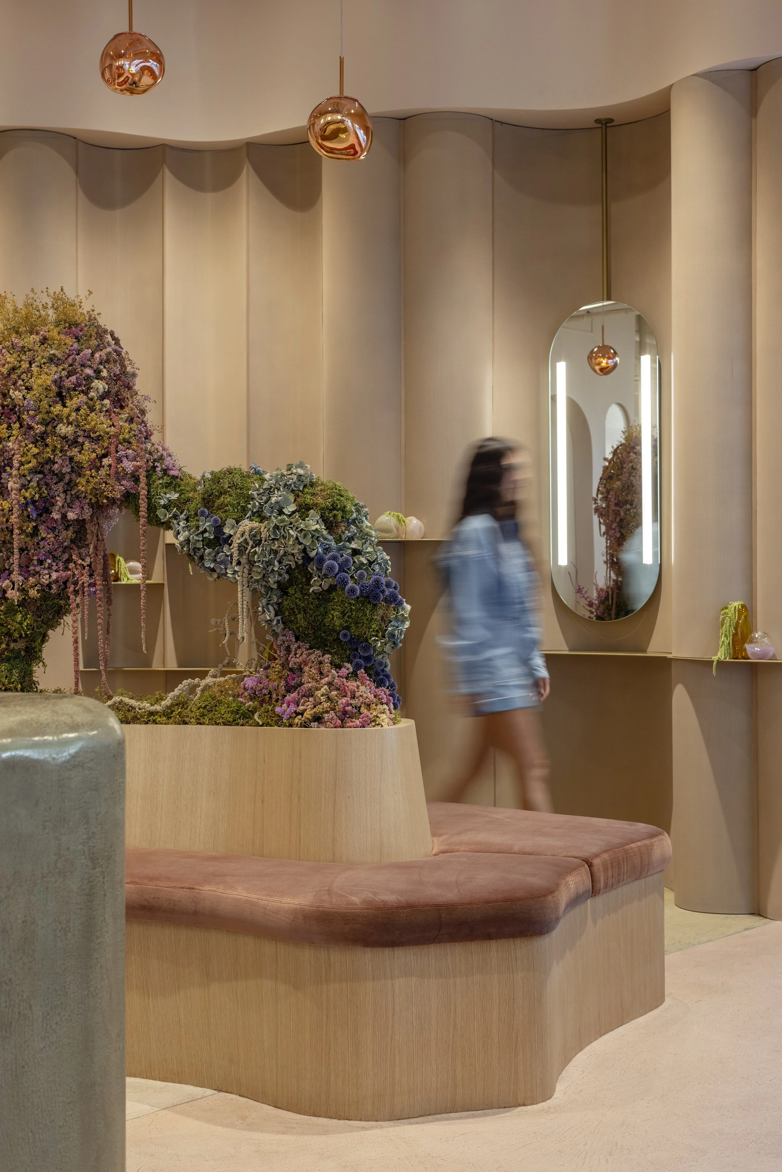 A woman walking past a curved, light wood bench with pink cushions, located in a modern interior space. The background includes a large mirror, a beige wall with vertical panels, pendant lights, and a floral arrangement with purple, pink, and green plants.