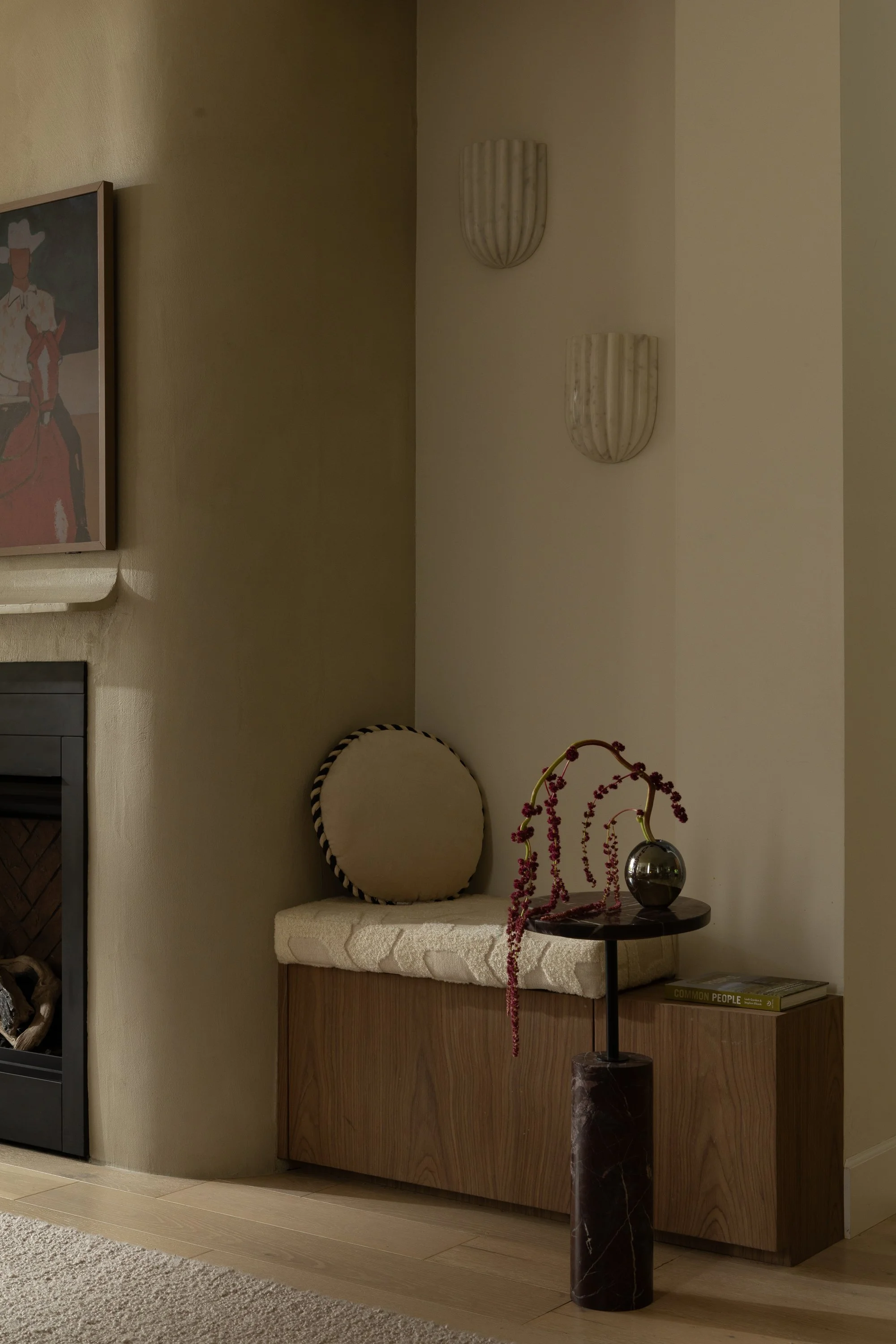 A cozy corner with a wooden cabinet topped with a white tufted cushion, a round cream pillow, a small black side table with a marble-textured black and gold vase containing pink beaded branches, and a book titled 'Common People'. The wall features two textured wall sconces and a framed artwork of a person in a hat, with beige walls and hardwood flooring.