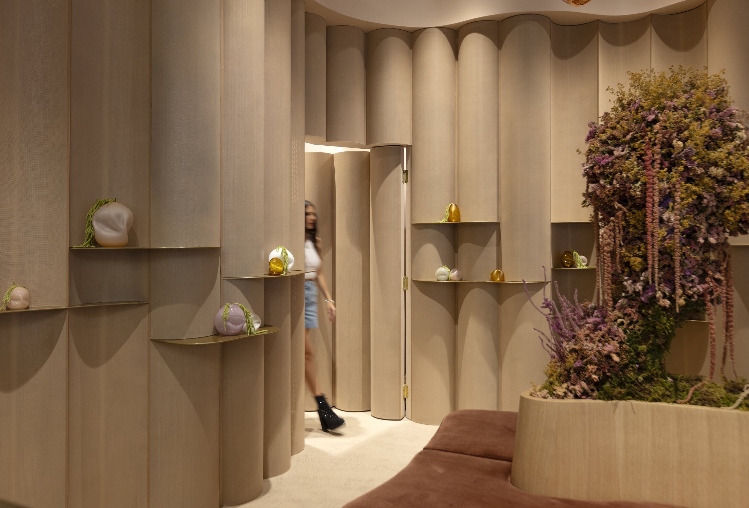 Interior view of a modern space with curved wooden wall panels decorated with various vases. A large floral arrangement is visible on the right side of the image. A woman is partially visible, walking through a door at the center of the scene.