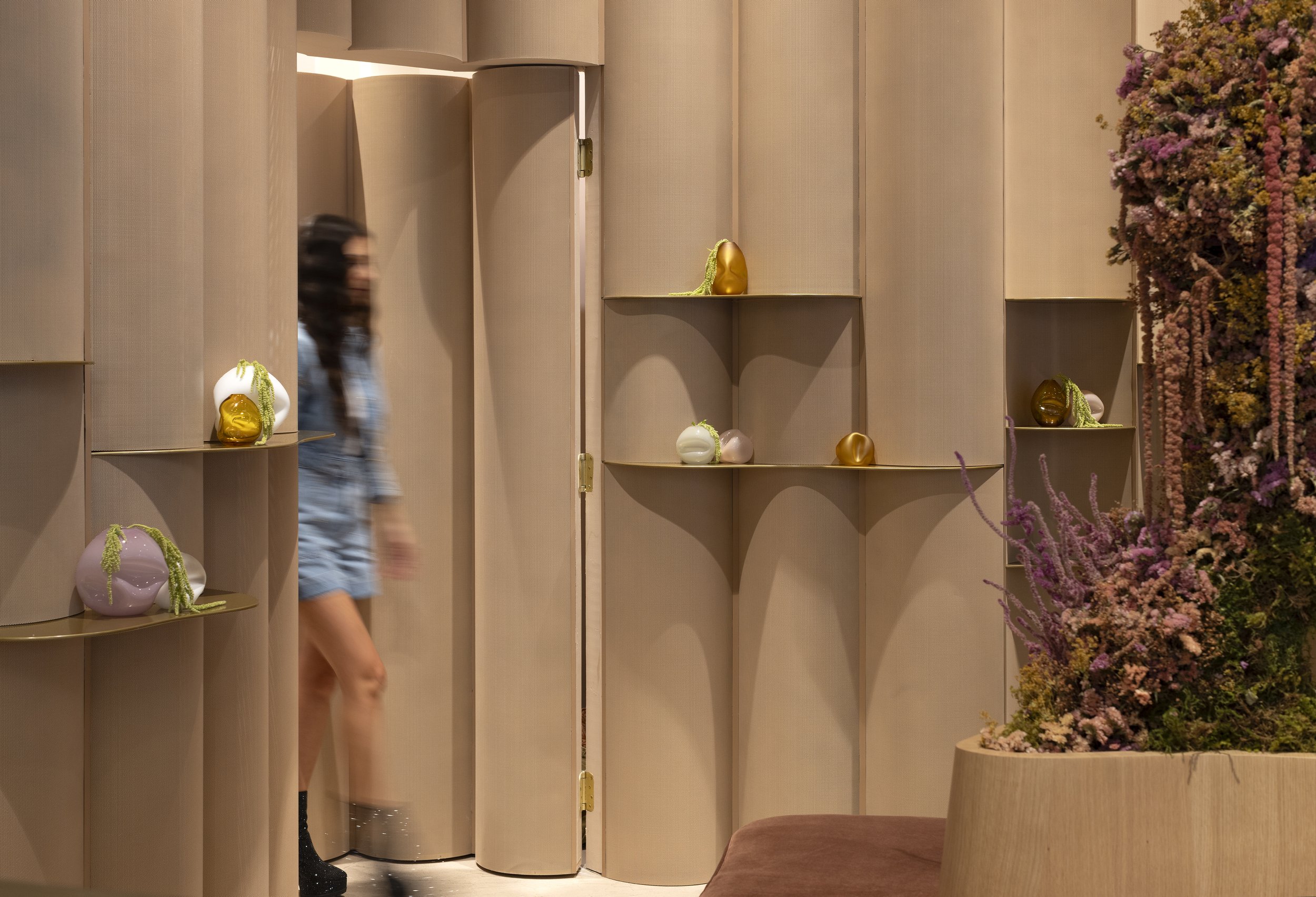 A woman walking past decorative shelves with vases, a closed beige door, and a large potted plant with pink and purple flowers.