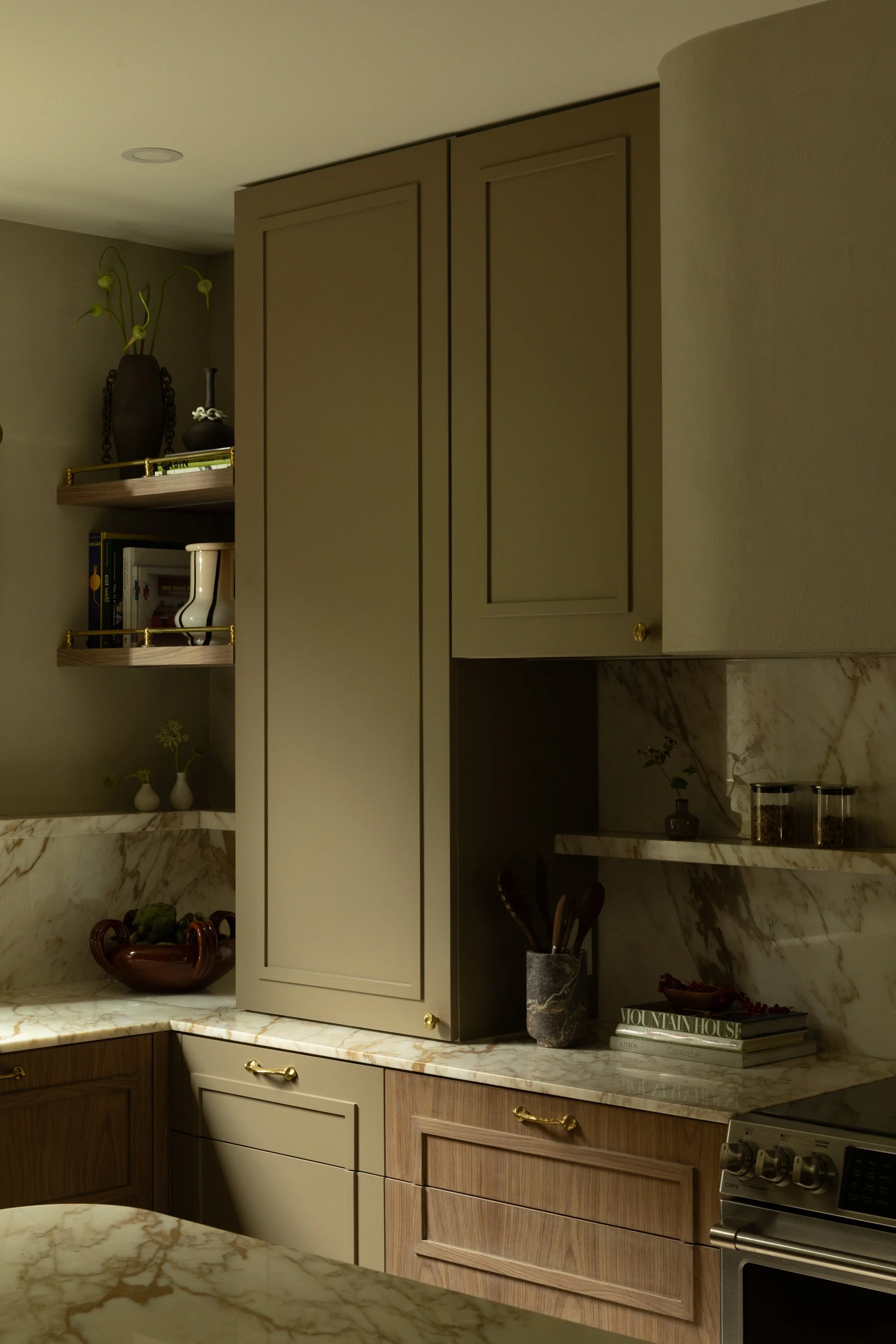 A kitchen with beige and brown cabinets, marble countertops, and open shelving with decorative items and books.