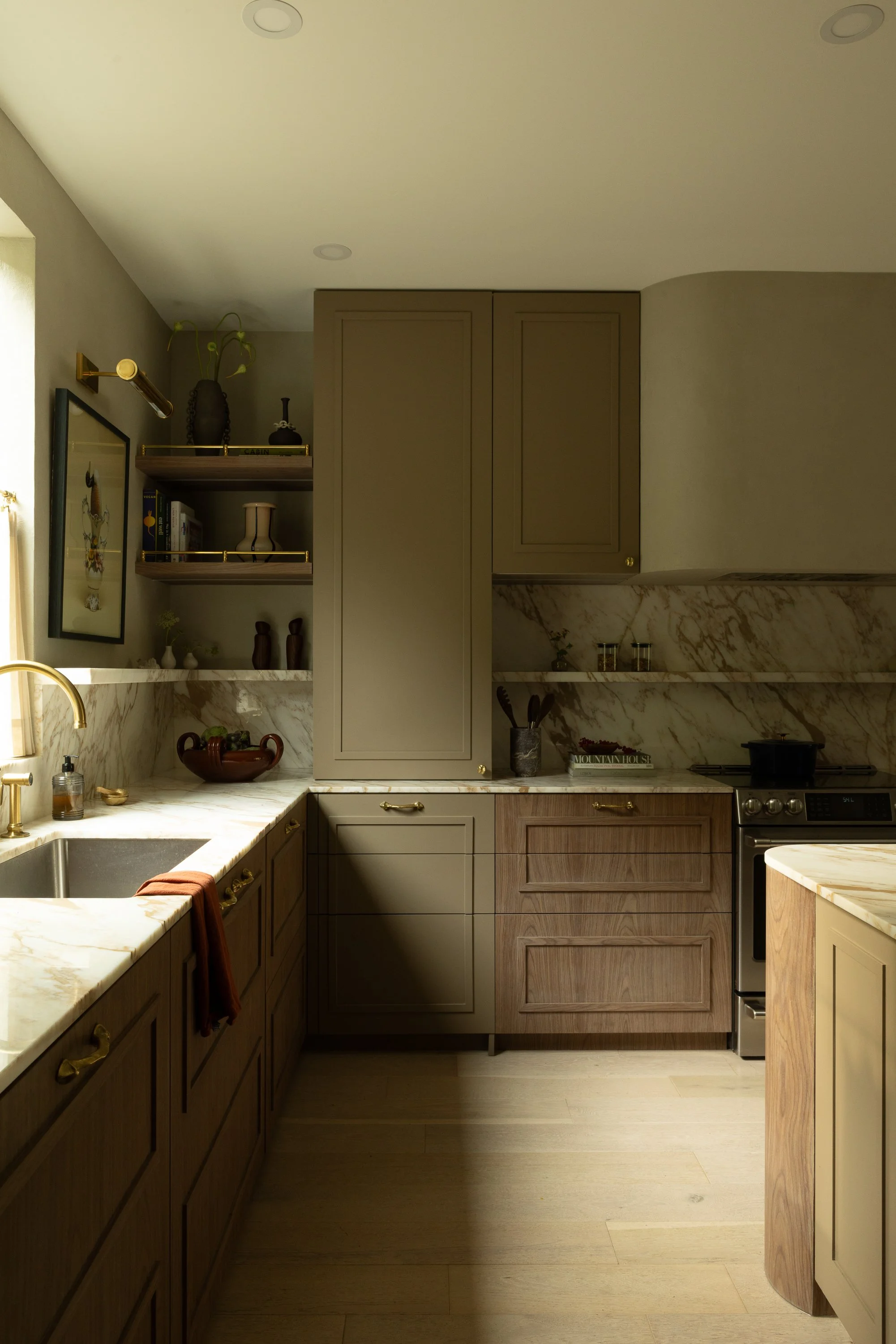 A kitchen with beige and brown cabinetry, marble countertops, open shelves with books and decorative items, a window with a curtain, and stainless steel appliances.