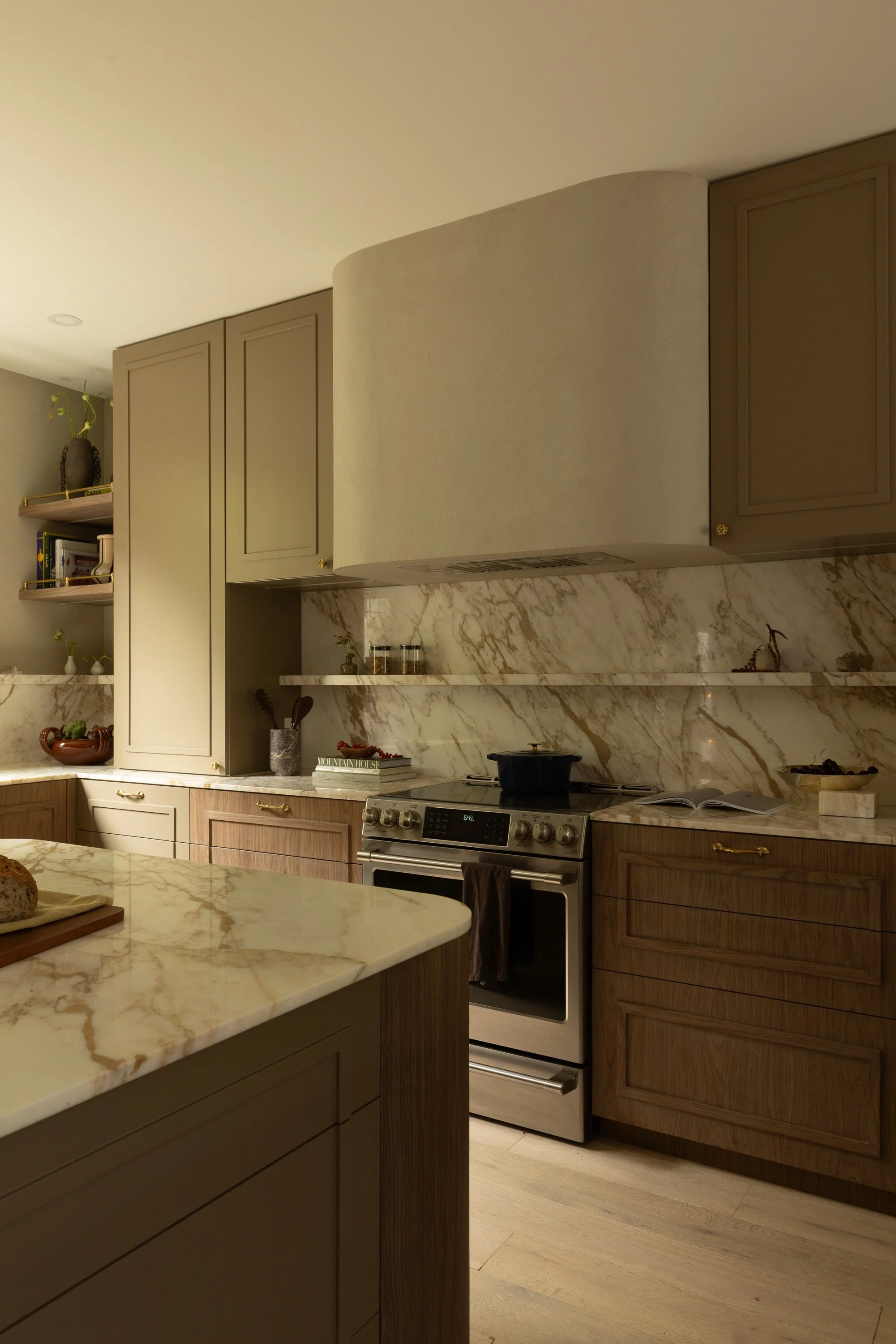 Modern kitchen with beige and wooden cabinets, marble countertops, stove, and open shelves with books and vases.