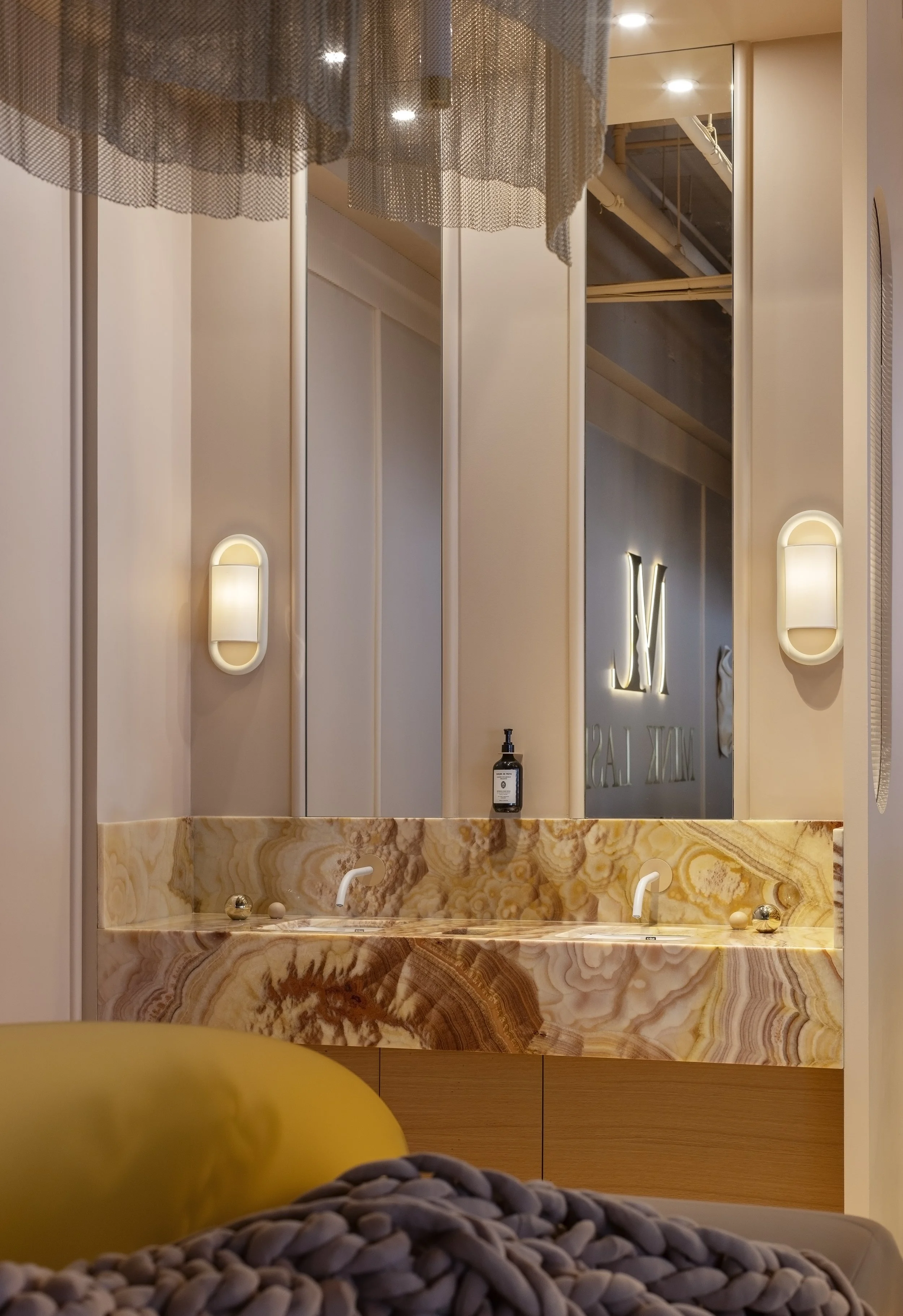 A bathroom vanity with a marble countertop and a mirrored wall with decorative lighting. Some objects and a blurred yellow item are in the foreground.