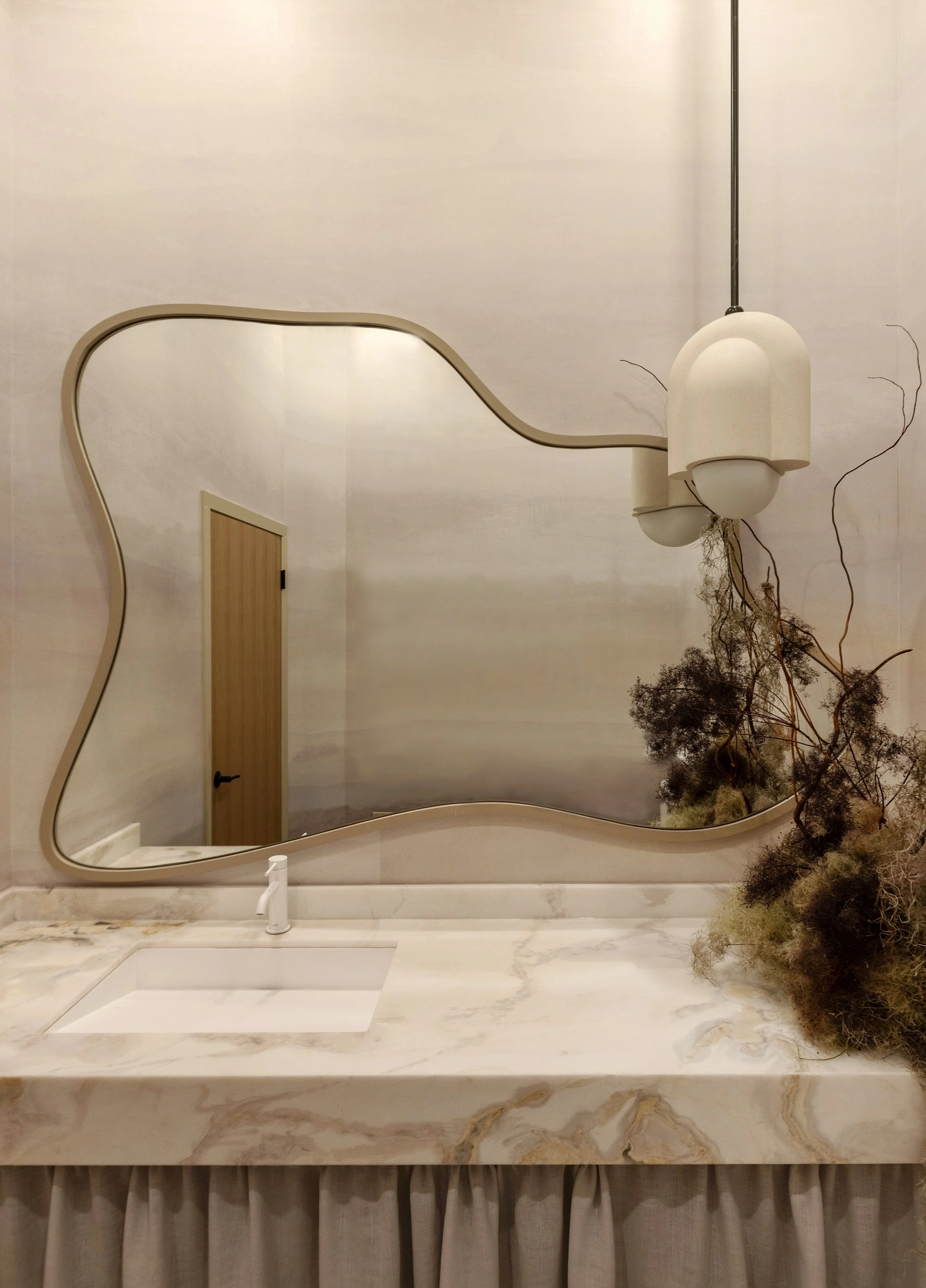 Modern bathroom with a marble countertop, a white sink, and a large irregularly shaped mirror. A unique hanging light fixture with three white globes and decorative dried plants are also visible.