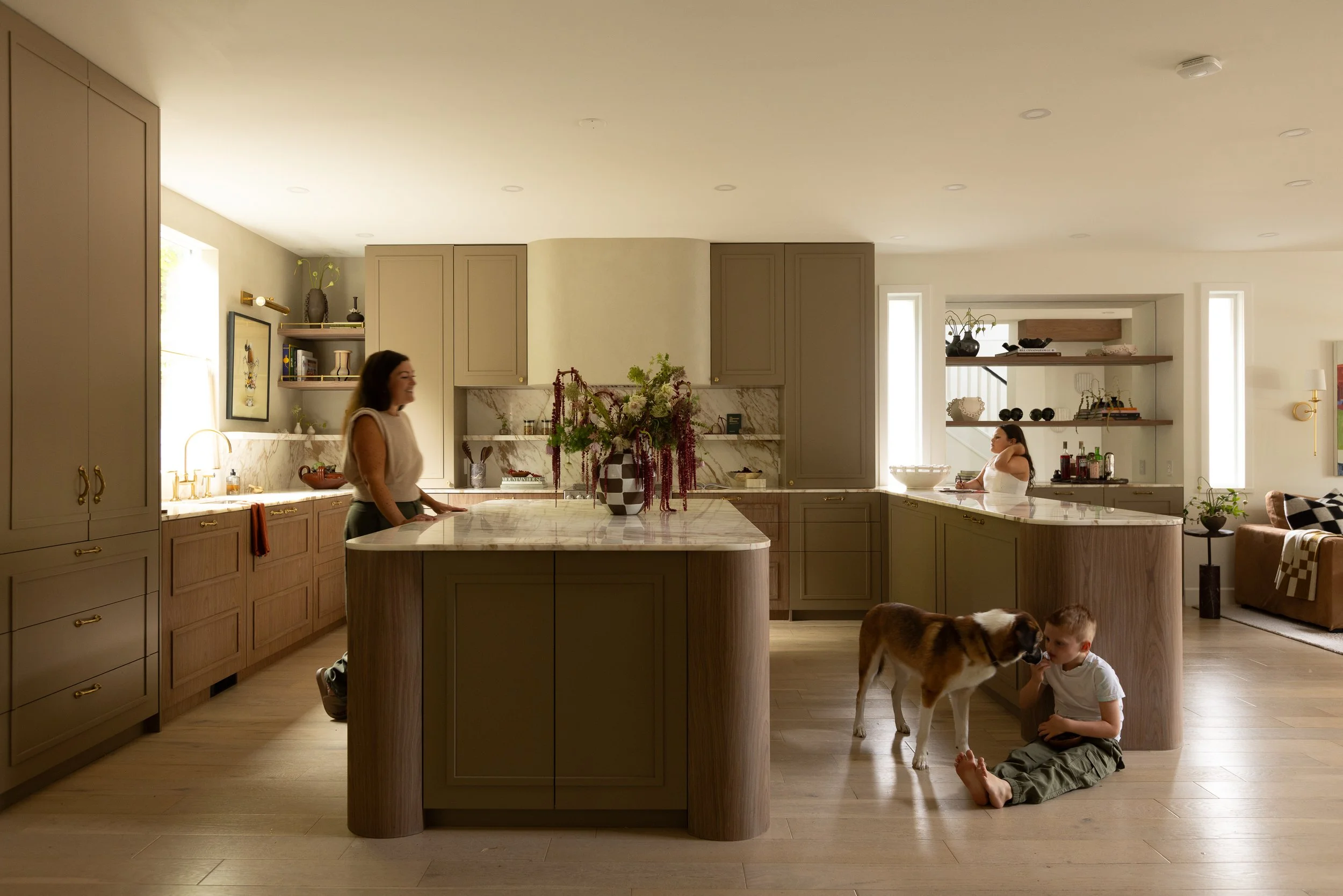 A modern kitchen with a large island, light-colored cabinetry, and open shelves filled with decorative items. Two women and two children are present; one woman is standing and smiling, while one child sits on the floor petting a dog, and the other child is sitting with the dog. The room is well-lit with natural light and has wooden flooring.