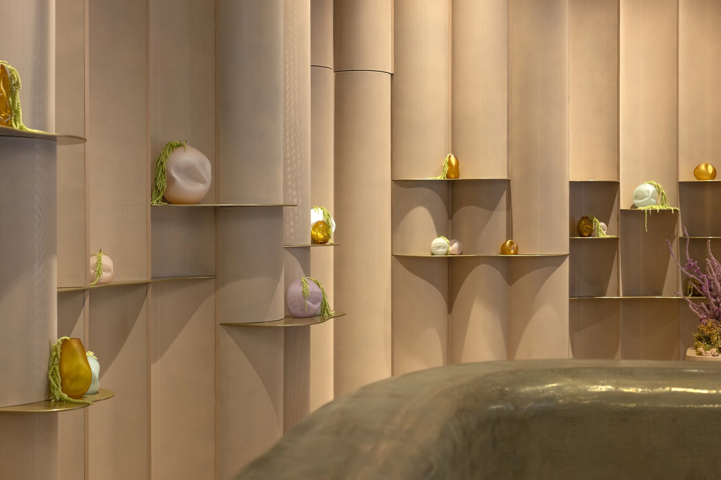 A wall display of colorful vases in various shapes and sizes, arranged on multiple small gold-colored shelves set into a beige, paneled wall.