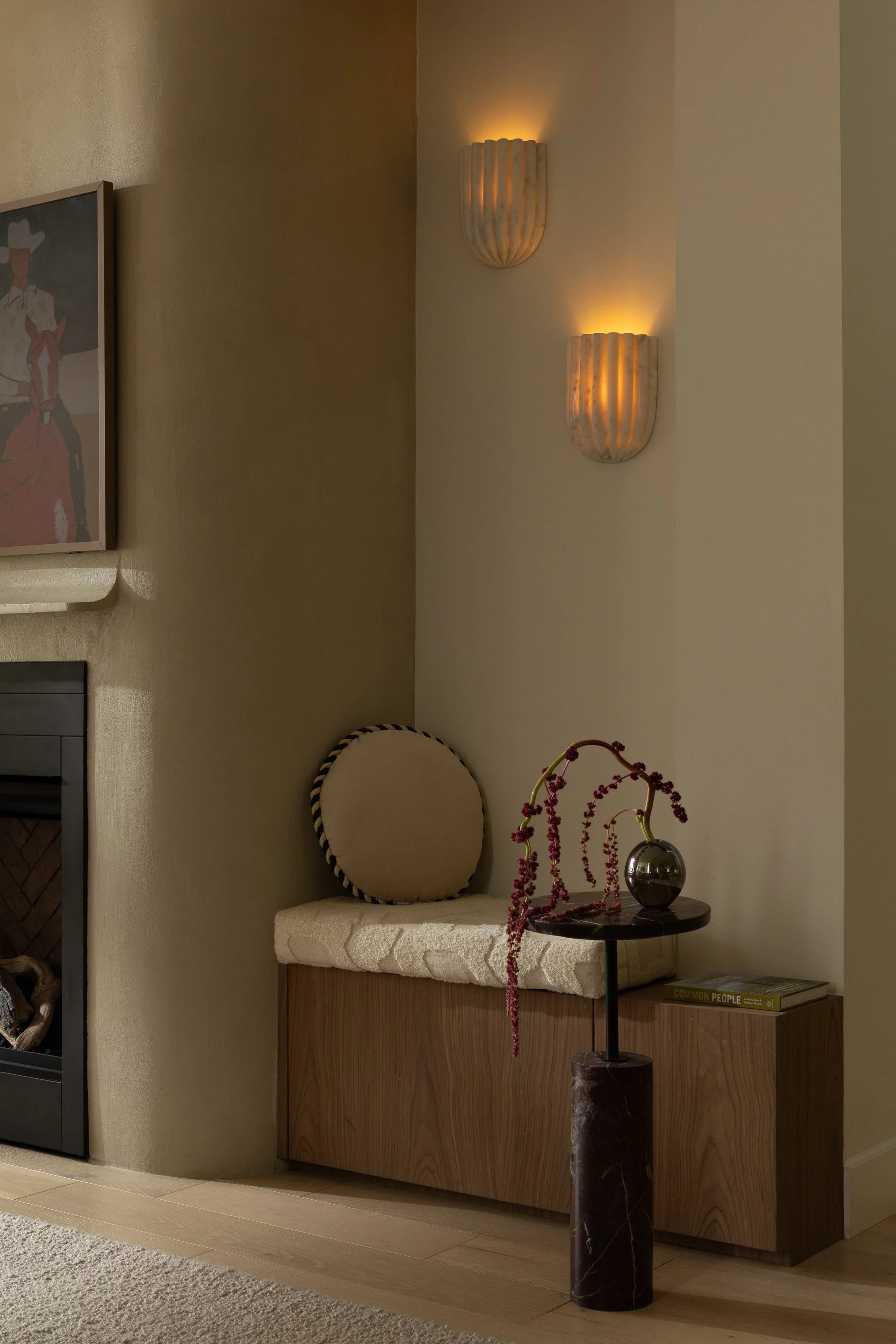 A corner of a living room with a wall-mounted light fixture, a wooden cabinet with a cushioned seat, a round pillow, and a side table with a vase of pinkish-red flowers, a black sculpture, and a book titled 'Common People'.