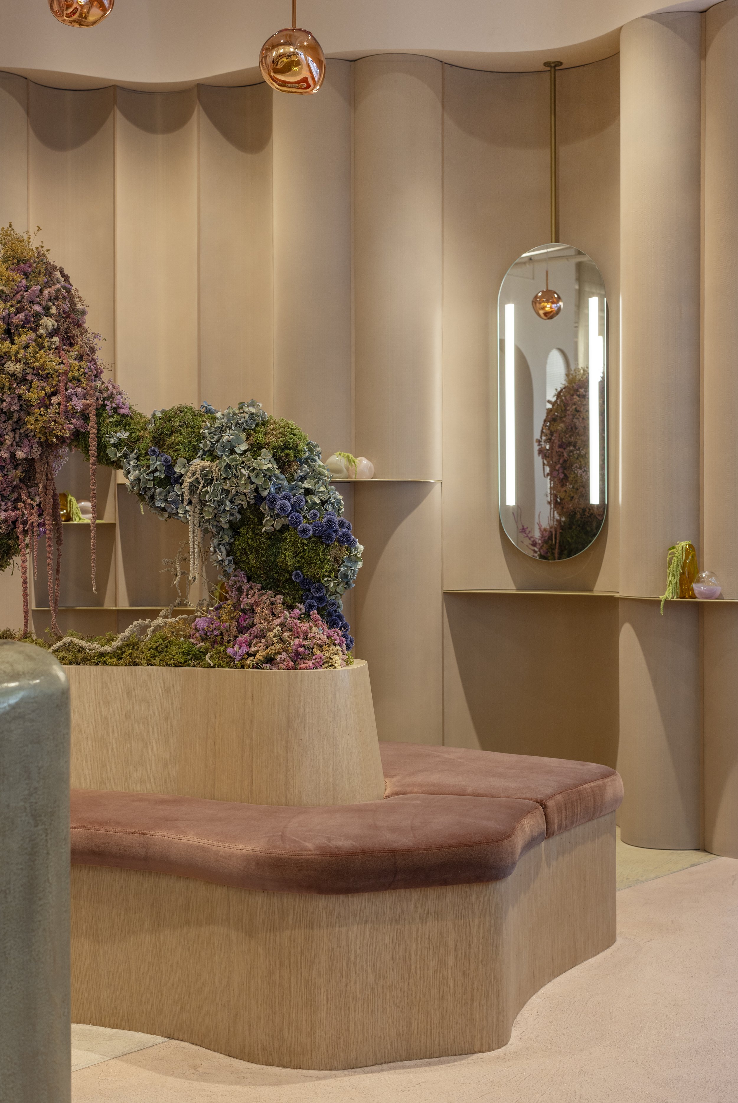 Interior of a modern space with a curved wooden seating area, large mirror, flower arrangement, hanging pendant lights, and subdued color palette.