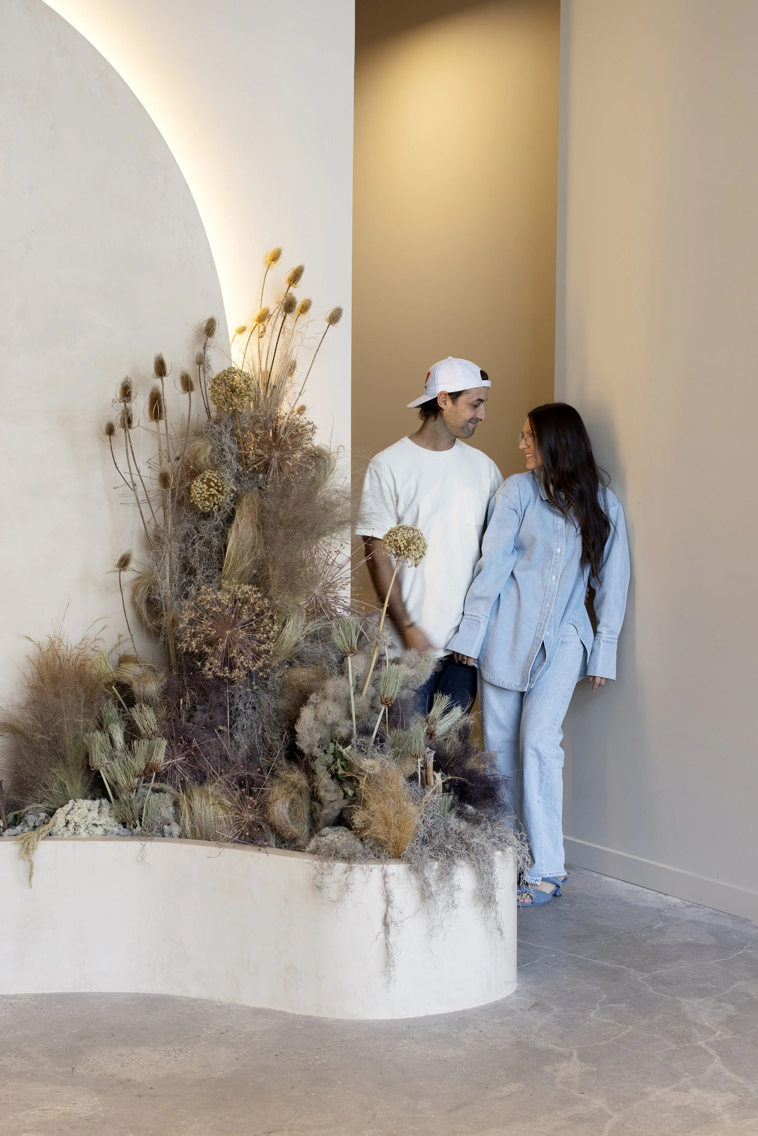 A man and woman are standing close together indoors, smiling at each other. The man is wearing a white t-shirt, a white cap worn backwards, and the woman is dressed in a light blue denim shirt and jeans. There is a large arrangement of dried plants and flowers on the left side of the image, with neutral-colored walls in the background.