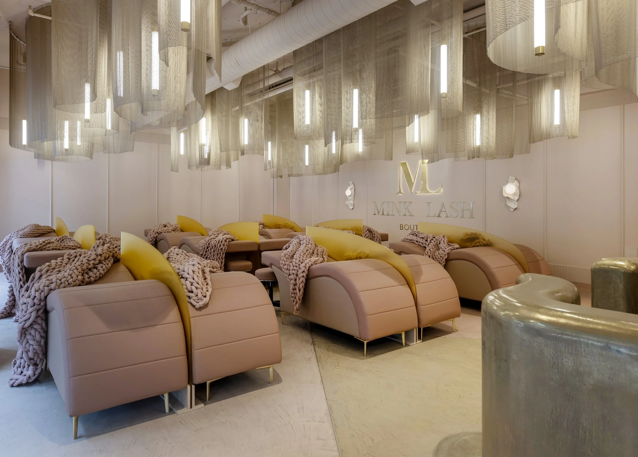 A stylish interior space featuring hanging fabric light fixtures, a wall with the logo 'MINK LASH,' plush pink seating with knitted blankets and yellow pillows, and a marble side table.