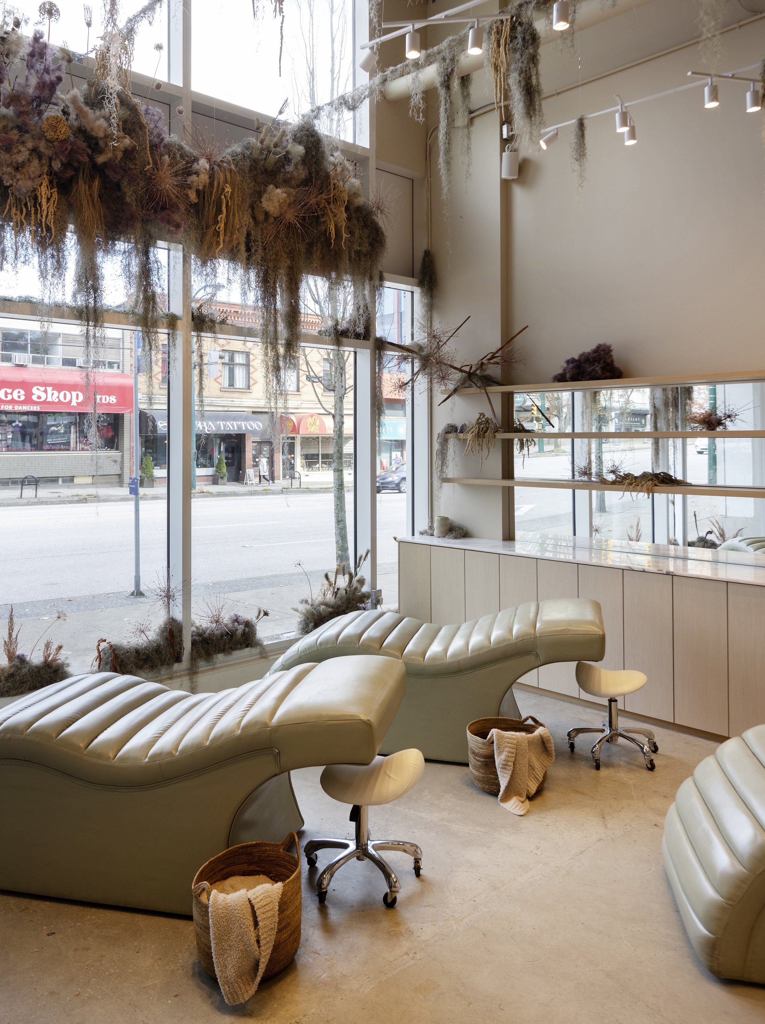 Interior of a modern spa or wellness center with two cream-colored, curvy lounge chairs and small stools. A large window showcases a city street with shops and pedestrians. Dried plants and mosses decorate the wall and window ledge, creating a natural, calming atmosphere.
