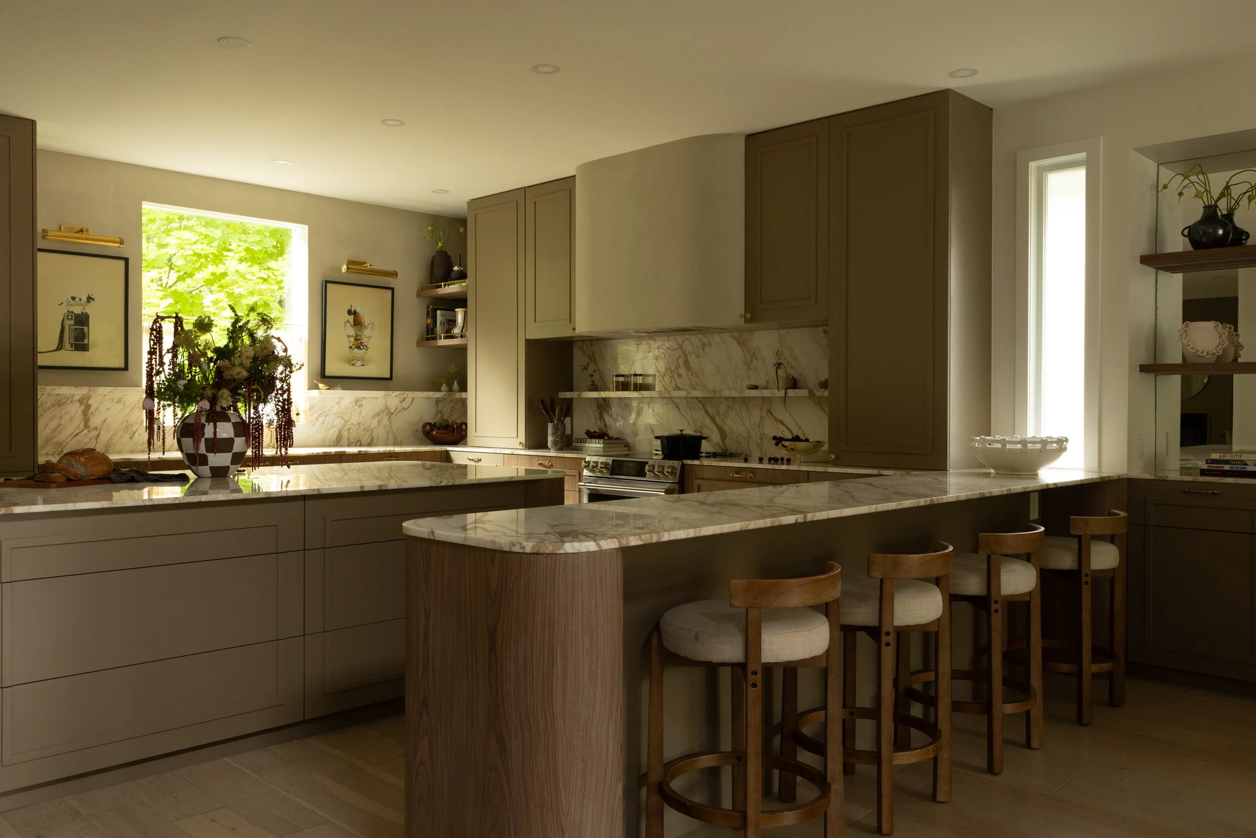 Modern kitchen with beige cabinets, marble countertops, bar seating, and artwork near a window with green foliage outside.