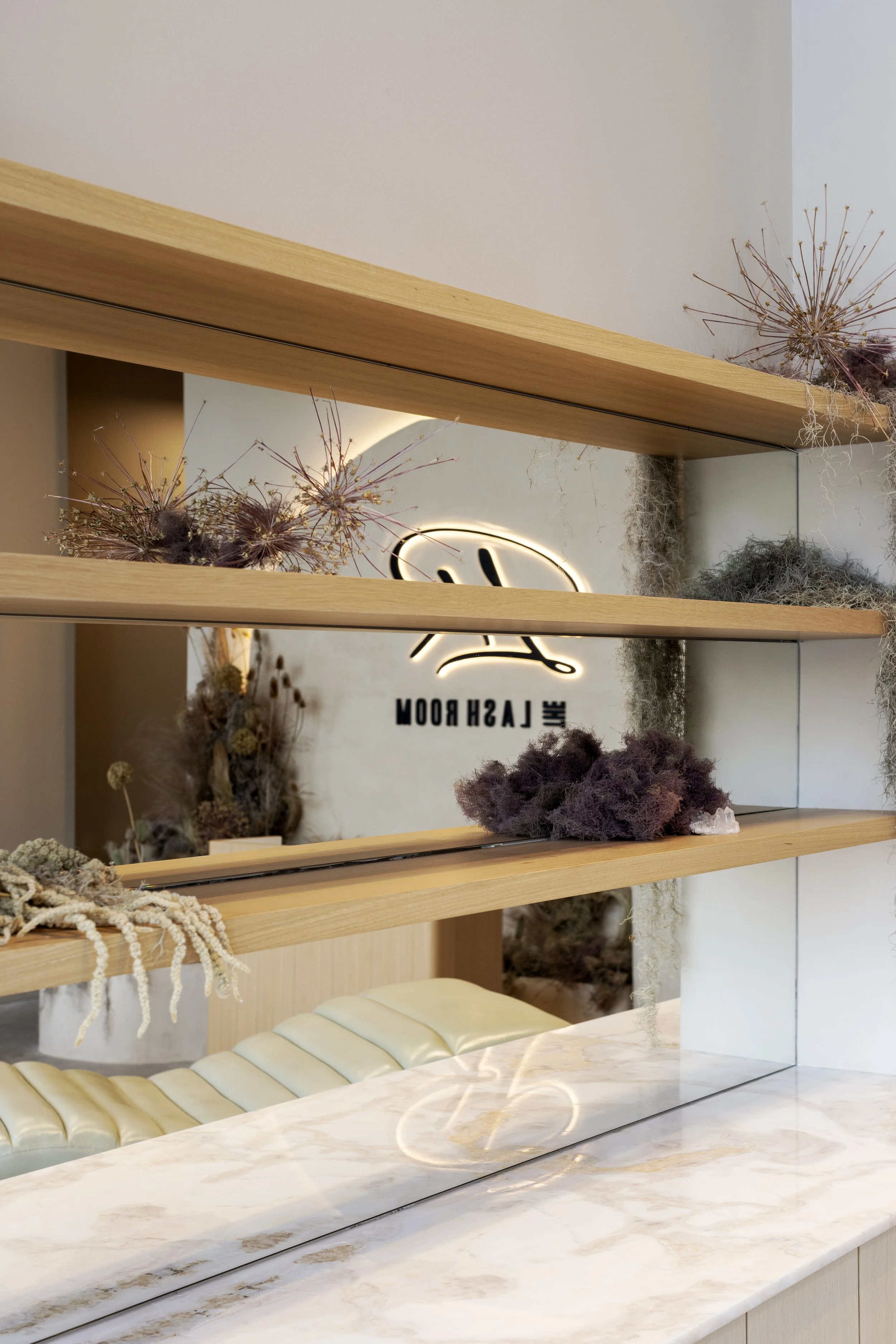 Decorative display shelf with dried plants, fluffy purple moss, and a neon sign in the background reading 'LAUGH ROOM' on a white wall, above a marble countertop.