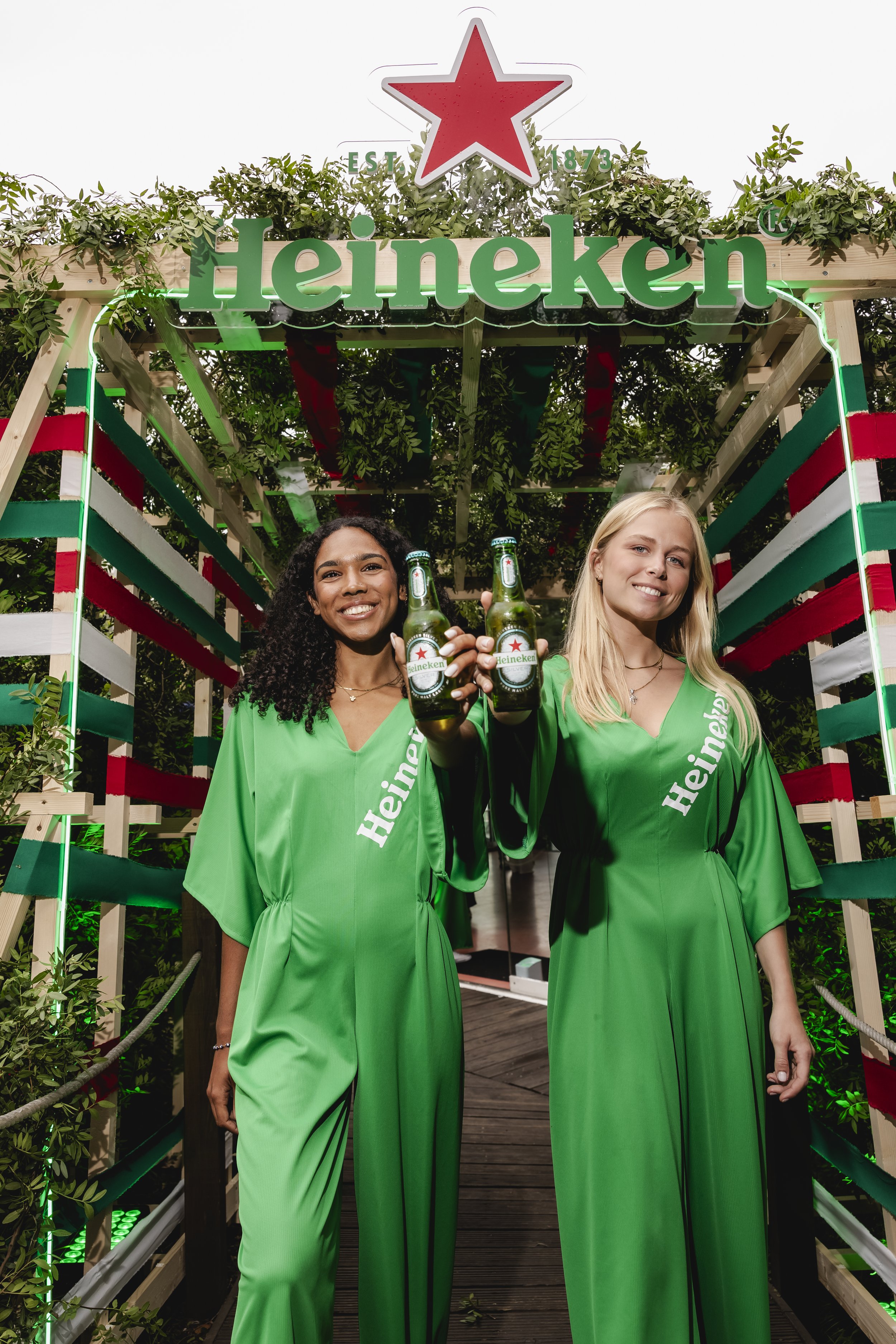Two women in green Heineken dresses holding beer bottles on a decorated outdoor walkway with Heineken signage and greenery overhead.