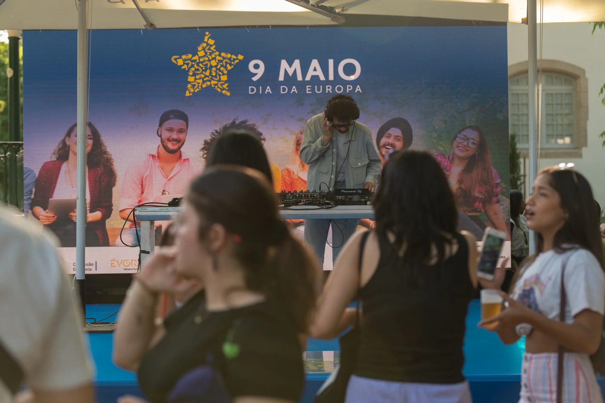 A DJ performing on stage with a large digital screen behind him displaying European Union's day celebration on May 9th, featuring a group of diverse people smiling. Audience members in the foreground watch the performance.