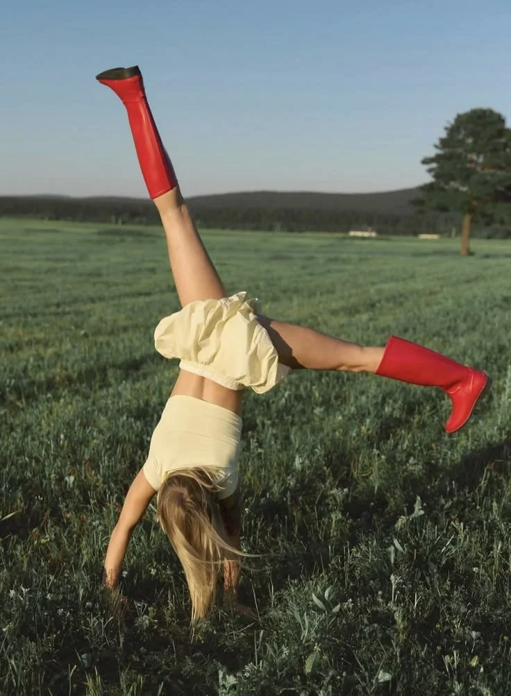 A girl doing a handstand in a grassy field, wearing a yellow top, cream-colored shorts, and red rain boots.