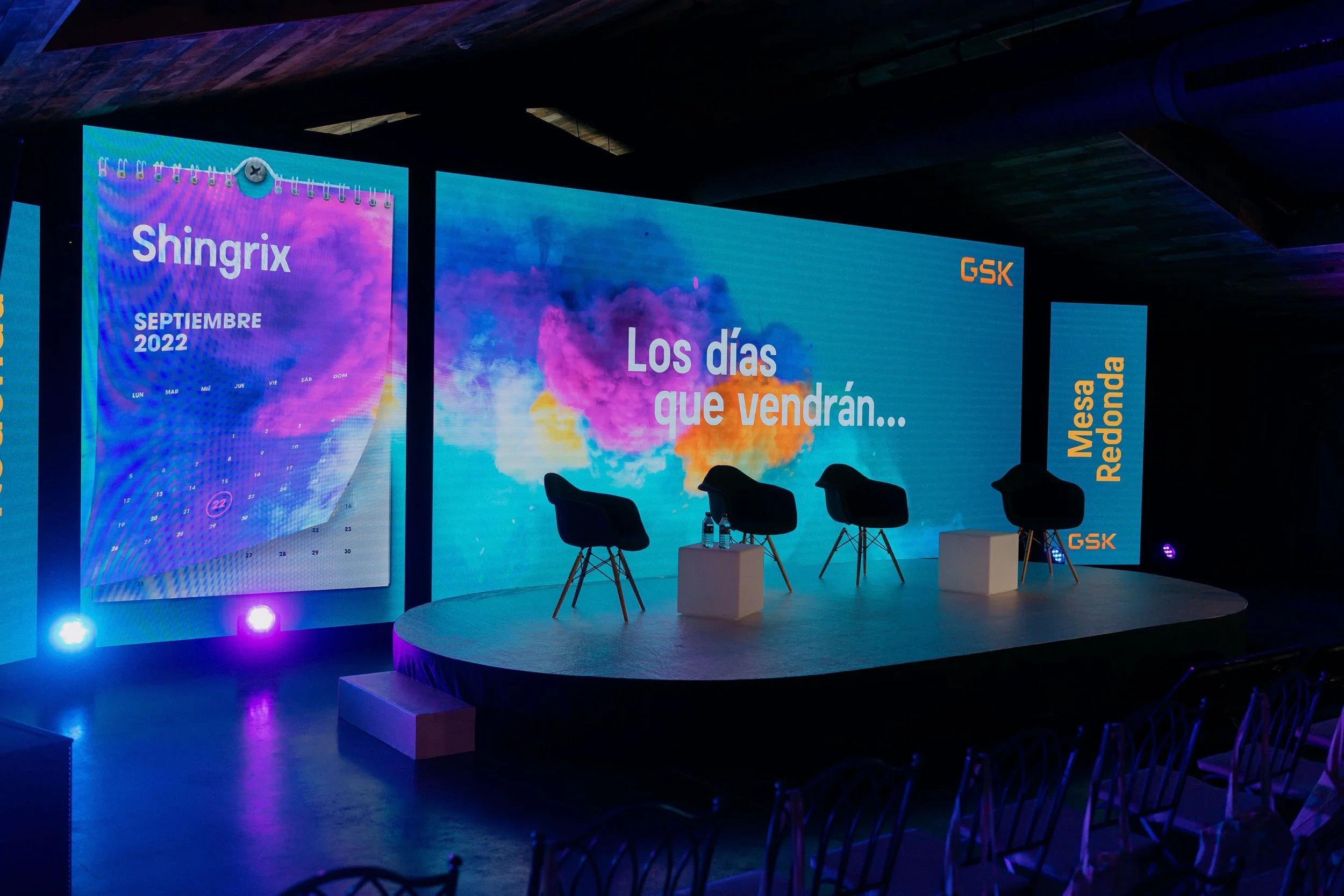 Large digital screen displays a calendar for September 2022 with colorful cloud background and the words "Los días que vendrán..." in Spanish, at an indoor event space with chairs and stage setup.