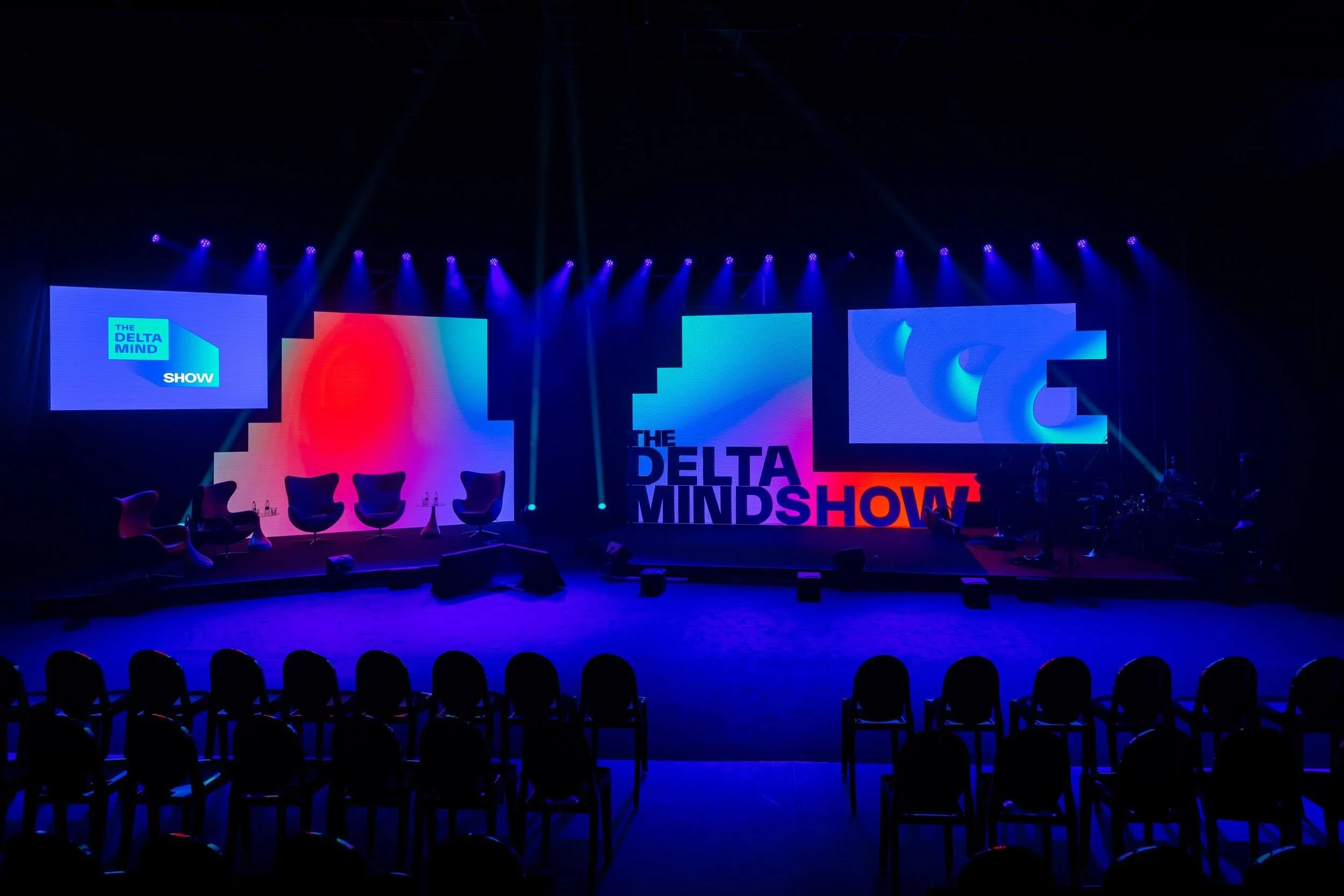 Stage setup with large LED screens displaying the logo and text 'The Delta Minds Show', multiple chairs arranged in a row, and blue stage lighting.