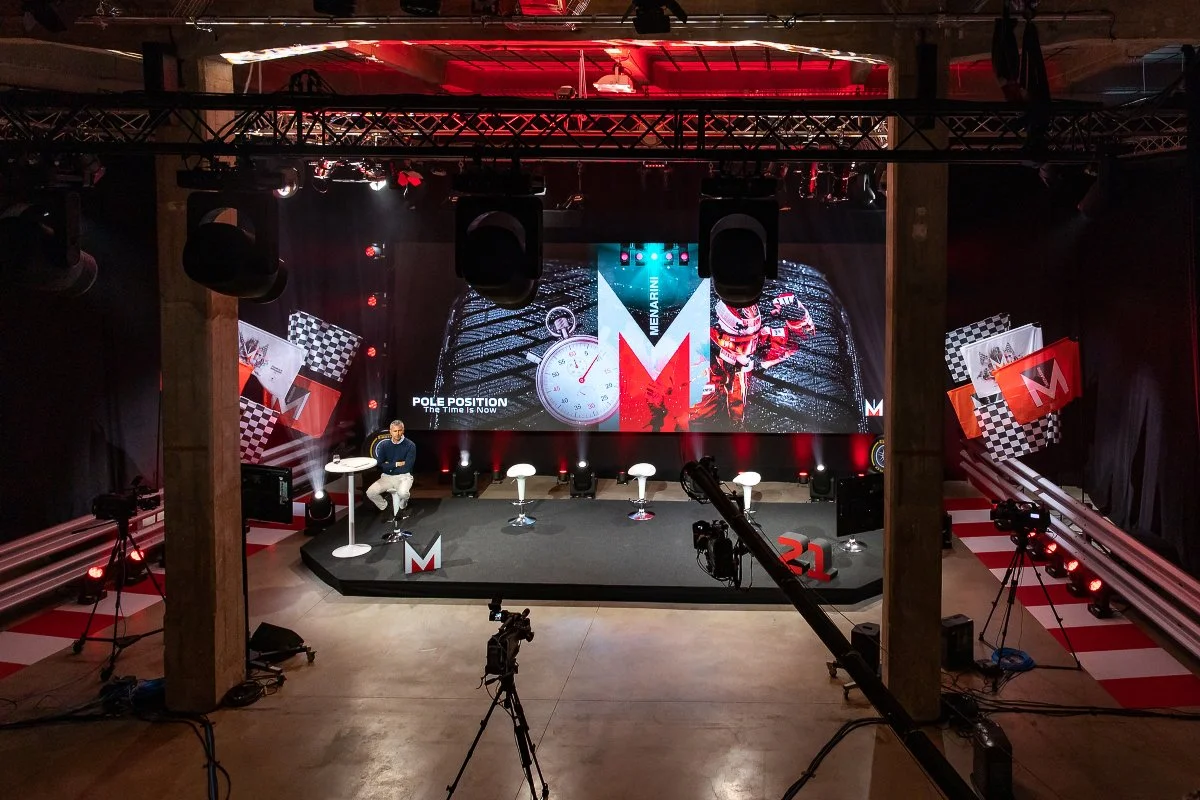 Stage set up for a presentation with a large screen displaying a watch, checkered flags, and racing scenes, decorated with red, black, and white elements, and several chairs and cameras.